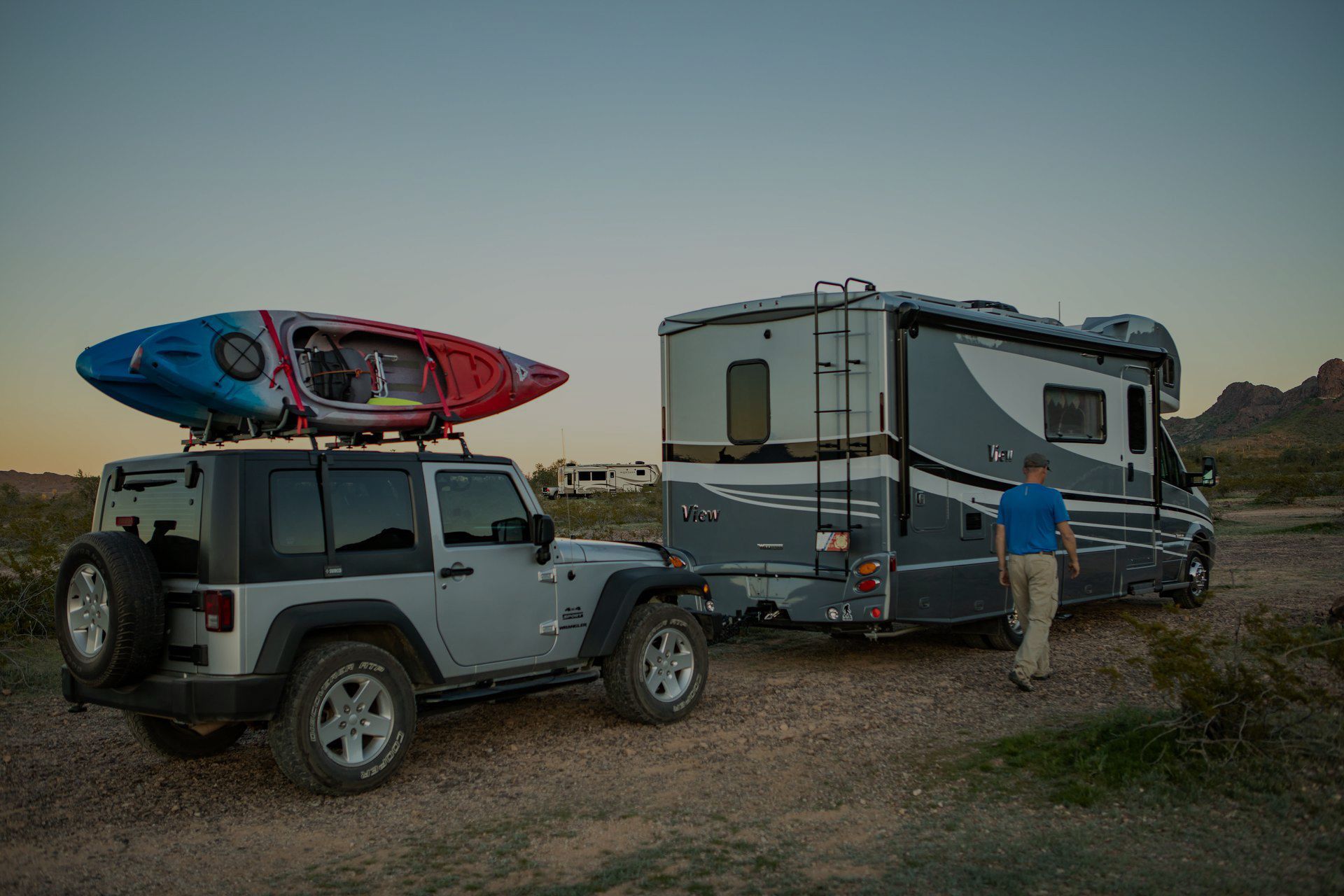 Gray RV and Jeep with kayak, parked in a desert setting