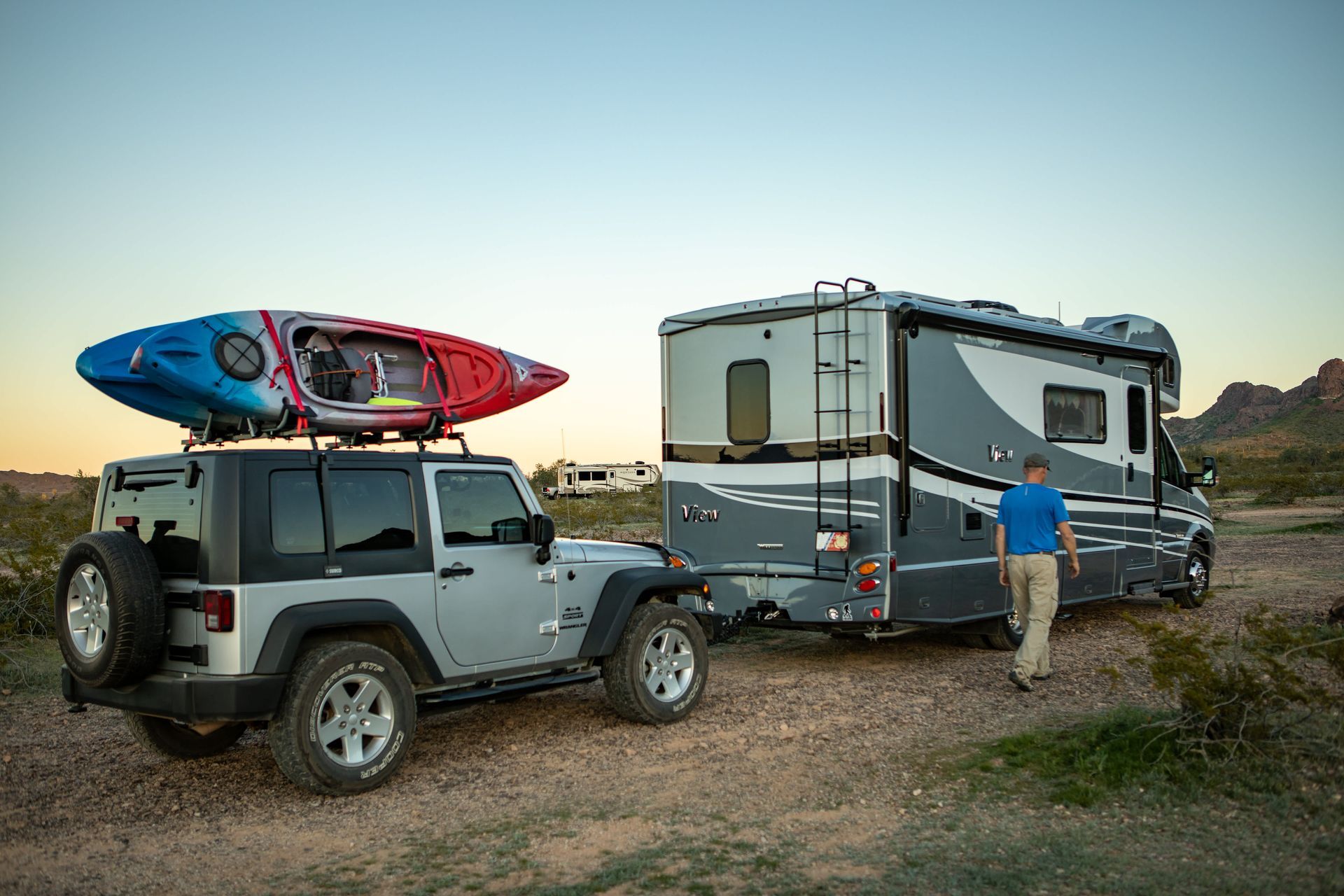 Gray RV and Jeep with kayak, parked in a desert setting