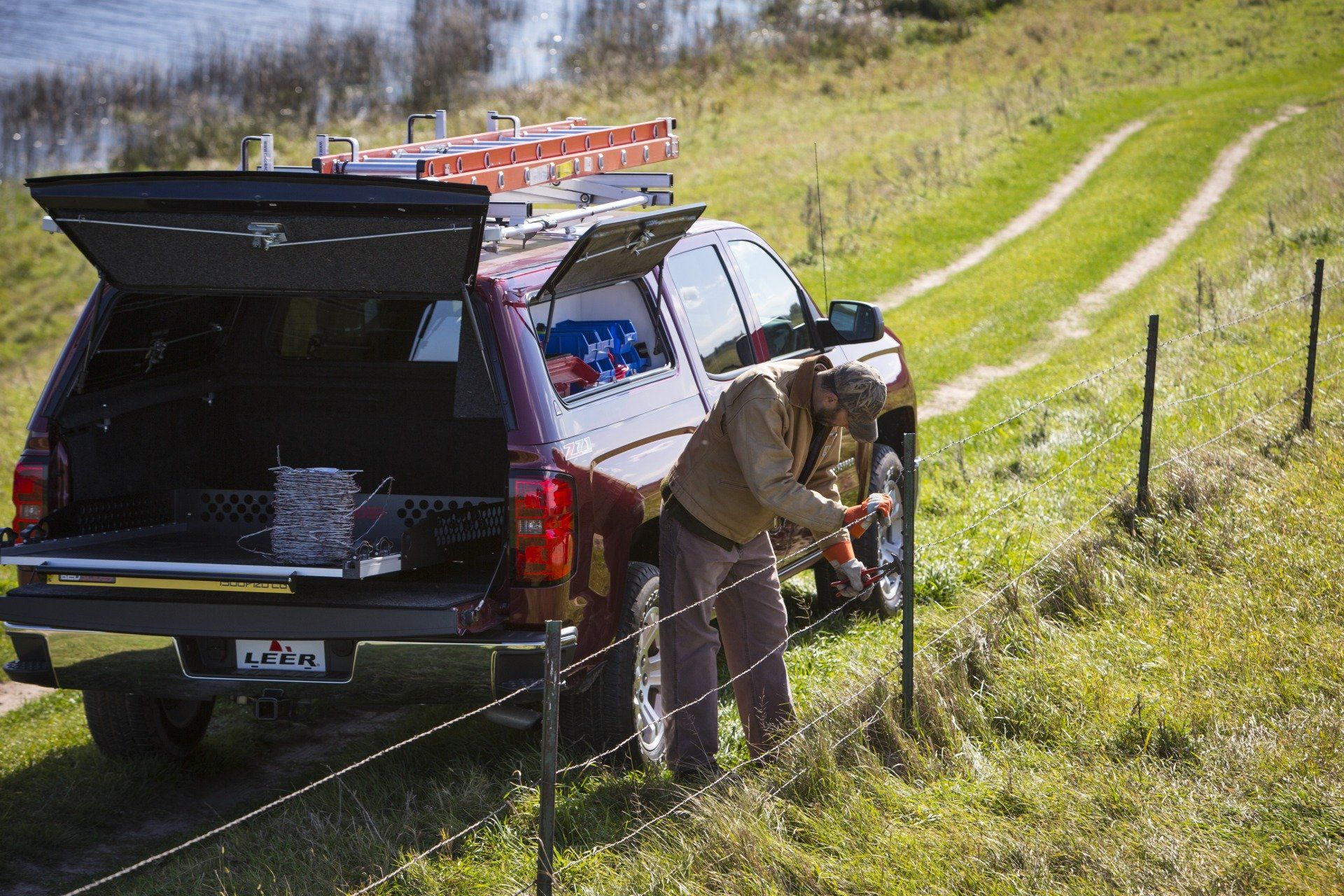 A man installs a fence post near a truck with an open bed, in a grassy field next to a body of water.