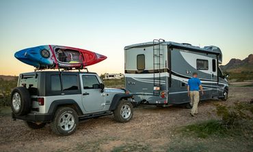 Silver Jeep with kayak towing a RV in a desert setting; man walks toard RV.