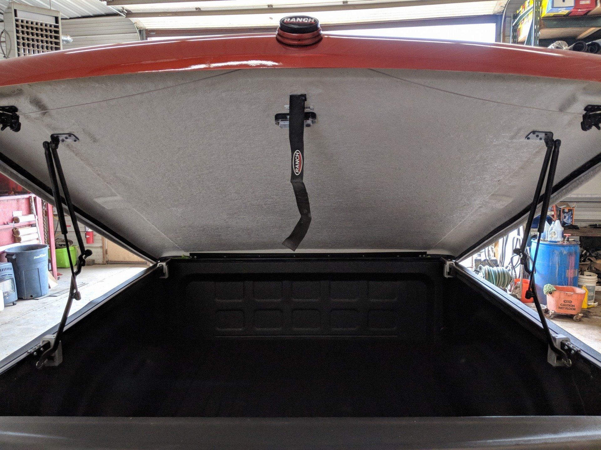 Open truck bed with red topper. Interior view of bed and topper hinges. A black strap holds the topper open.
