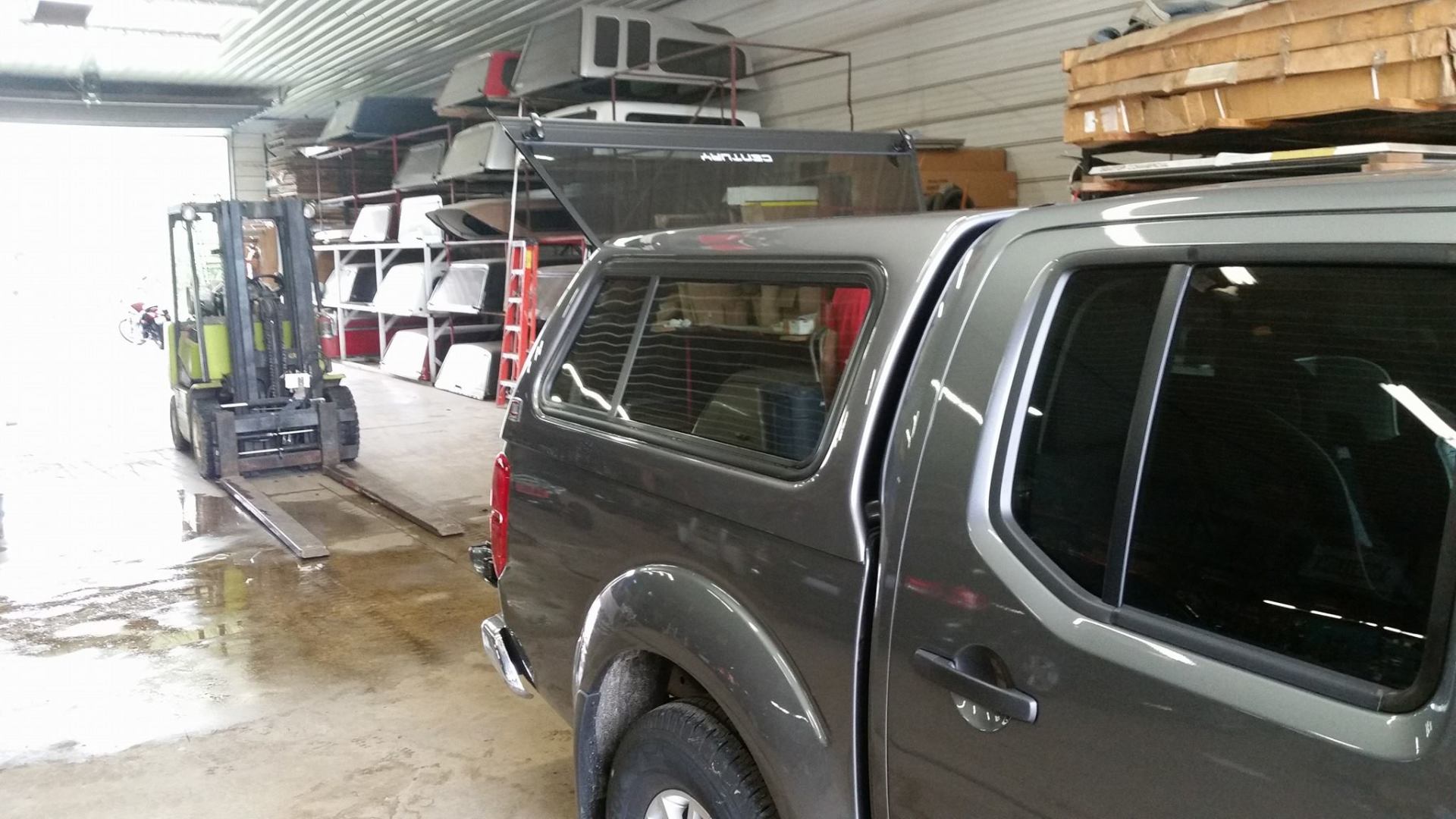 Truck with canopy next to forklift in a warehouse. Various truck canopies on shelves.