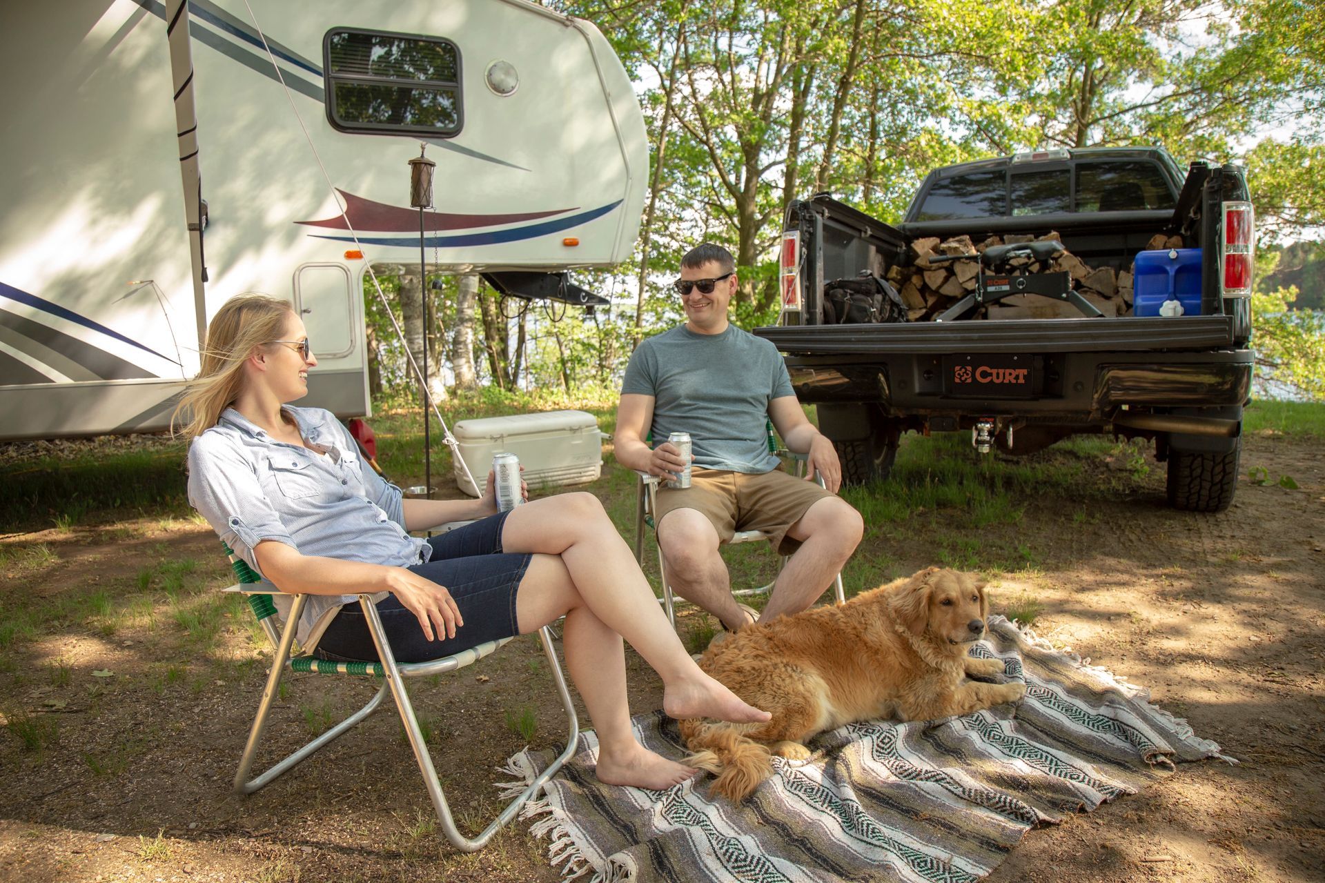 A couple relaxes outside an RV with a dog, a truck bed with firewood, and a cooler in nature.