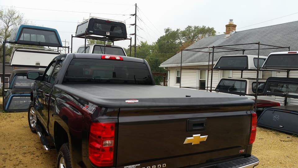 Black pickup truck with closed bed cover, surrounded by truck toppers. Outdoor setting.