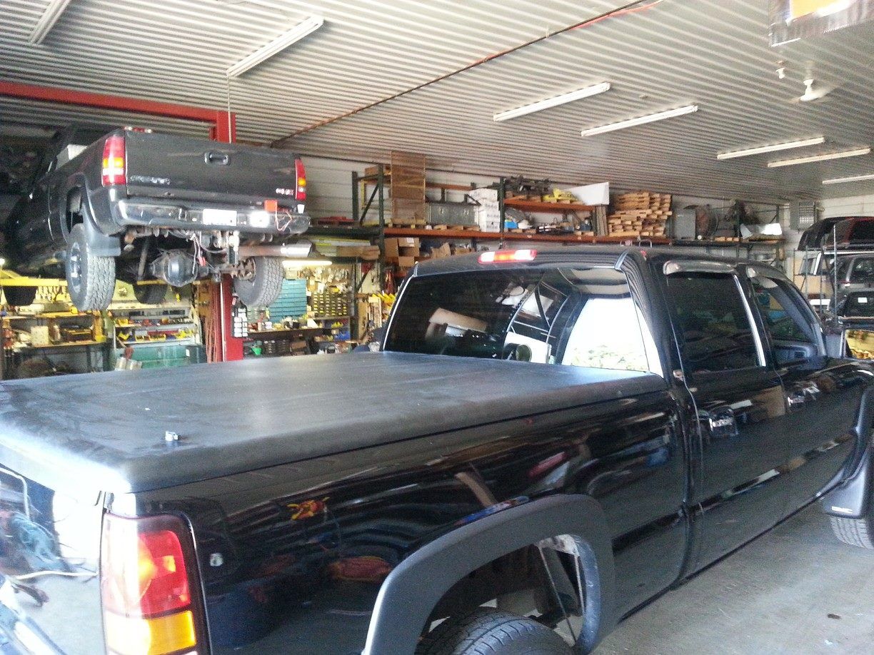 Black pickup truck in a garage; another truck on a lift in the background, surrounded by tools and equipment.