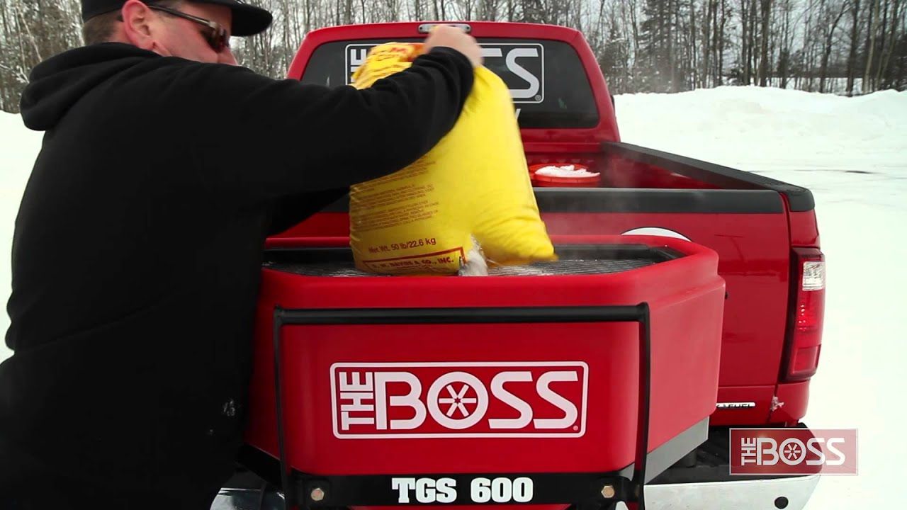 Person loading a yellow salt bag into a red Boss TGS 600 spreader on a red truck bed in snowy conditions.