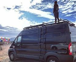Black van with ladder, person standing on the roof against a cloudy sky.