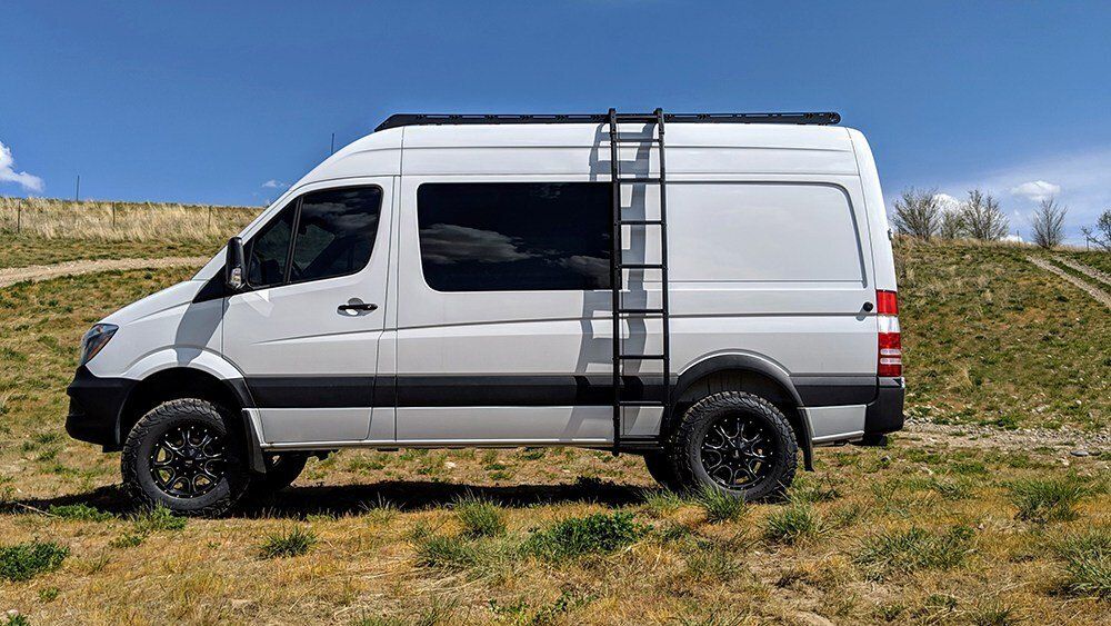 White van with a black ladder on its side, parked in a grassy area under a blue sky.