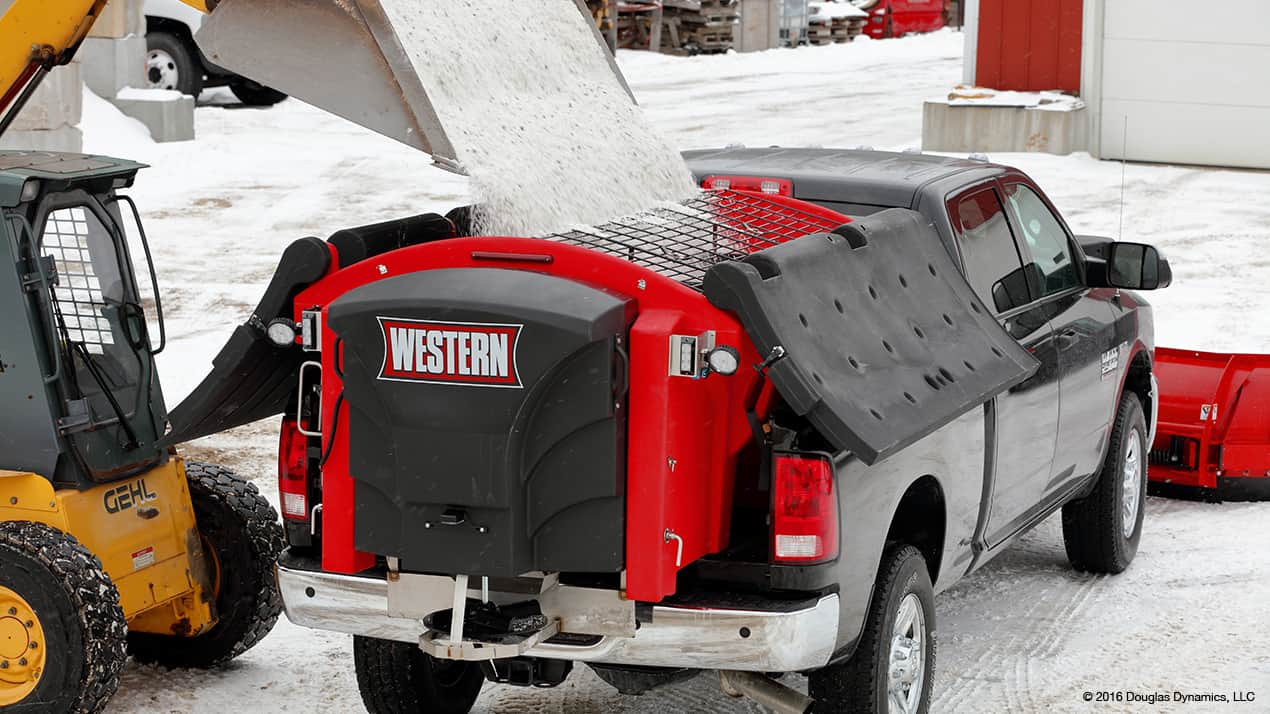 A truck is being loaded with material from a skid steer loader. A red and black snowplow attachment is mounted on the truck bed.