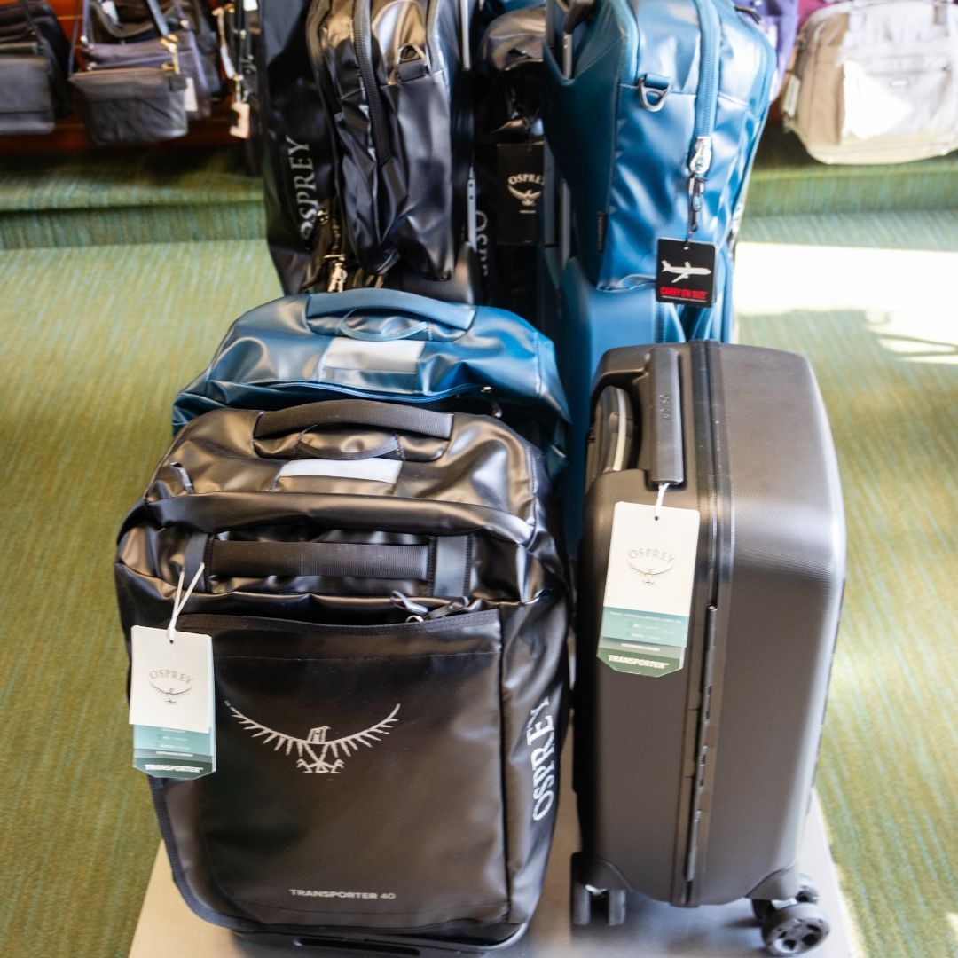 A display of Osprey brand rolling suitcases and backpacks in black, teal, and gray in a store with green carpet.
