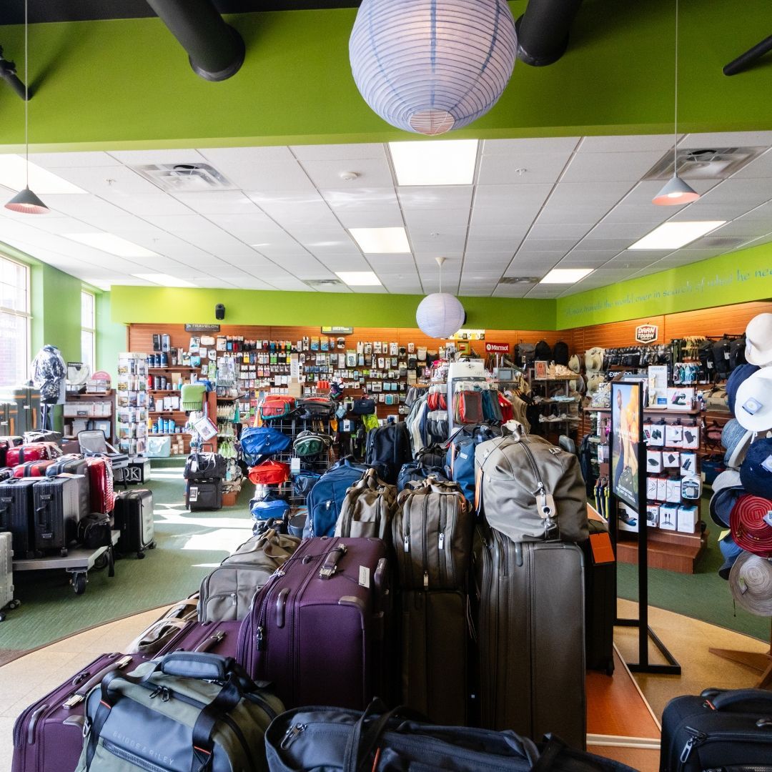 A retail store displays a wide variety of luggage, suitcases, and travel gear on shelves and floor racks under bright lights.