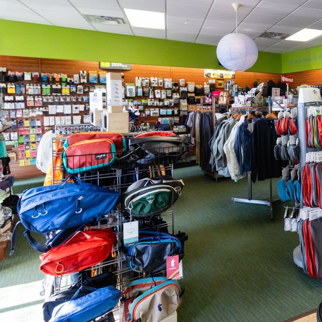 A retail store aisle featuring displays of colorful fanny packs, bags, and hanging clothing racks.