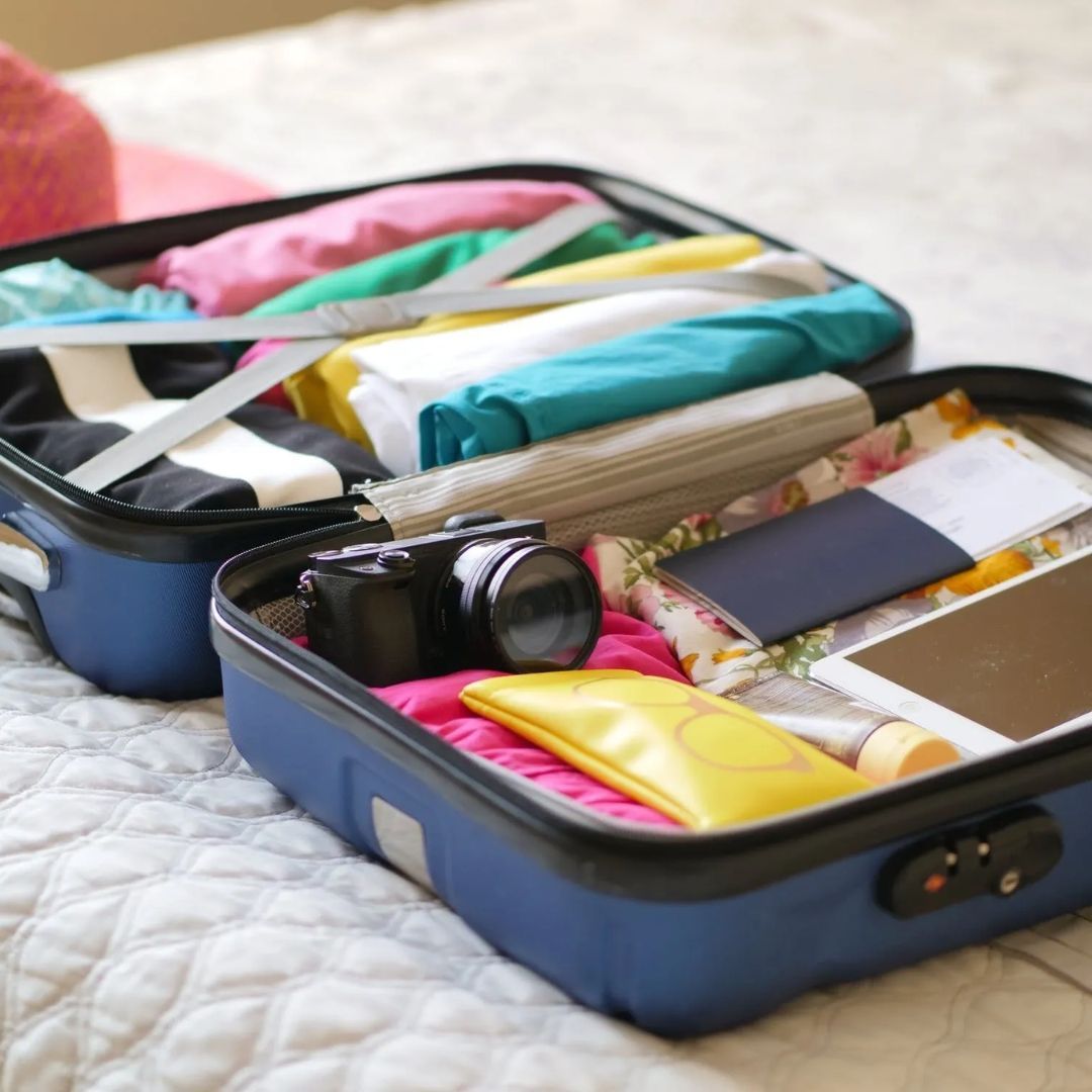 An open blue suitcase on a bed, packed with neatly folded clothes, a camera, a passport, and travel essentials.