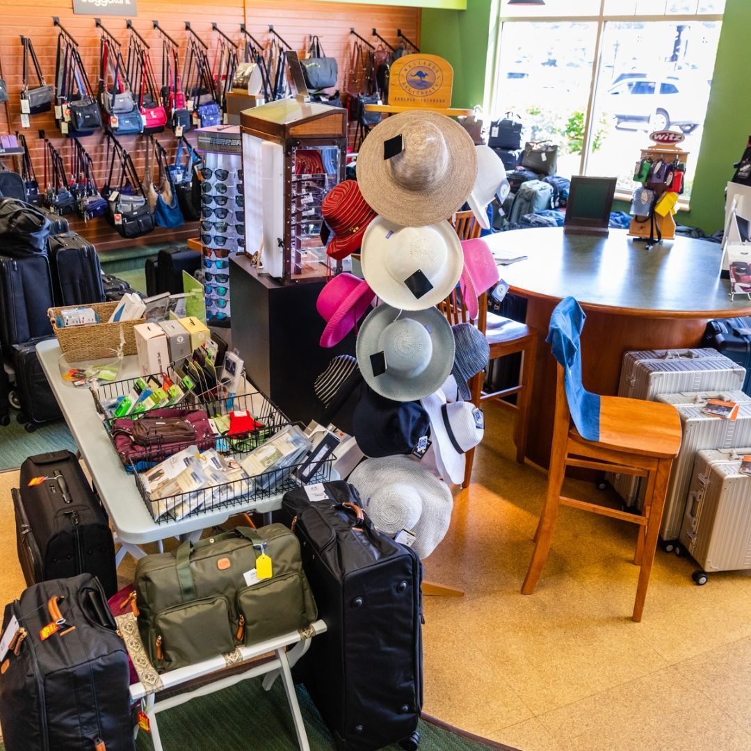 A shop interior with a hat display rack, a table of travel accessories, luggage, and a wooden table with a blue chair.