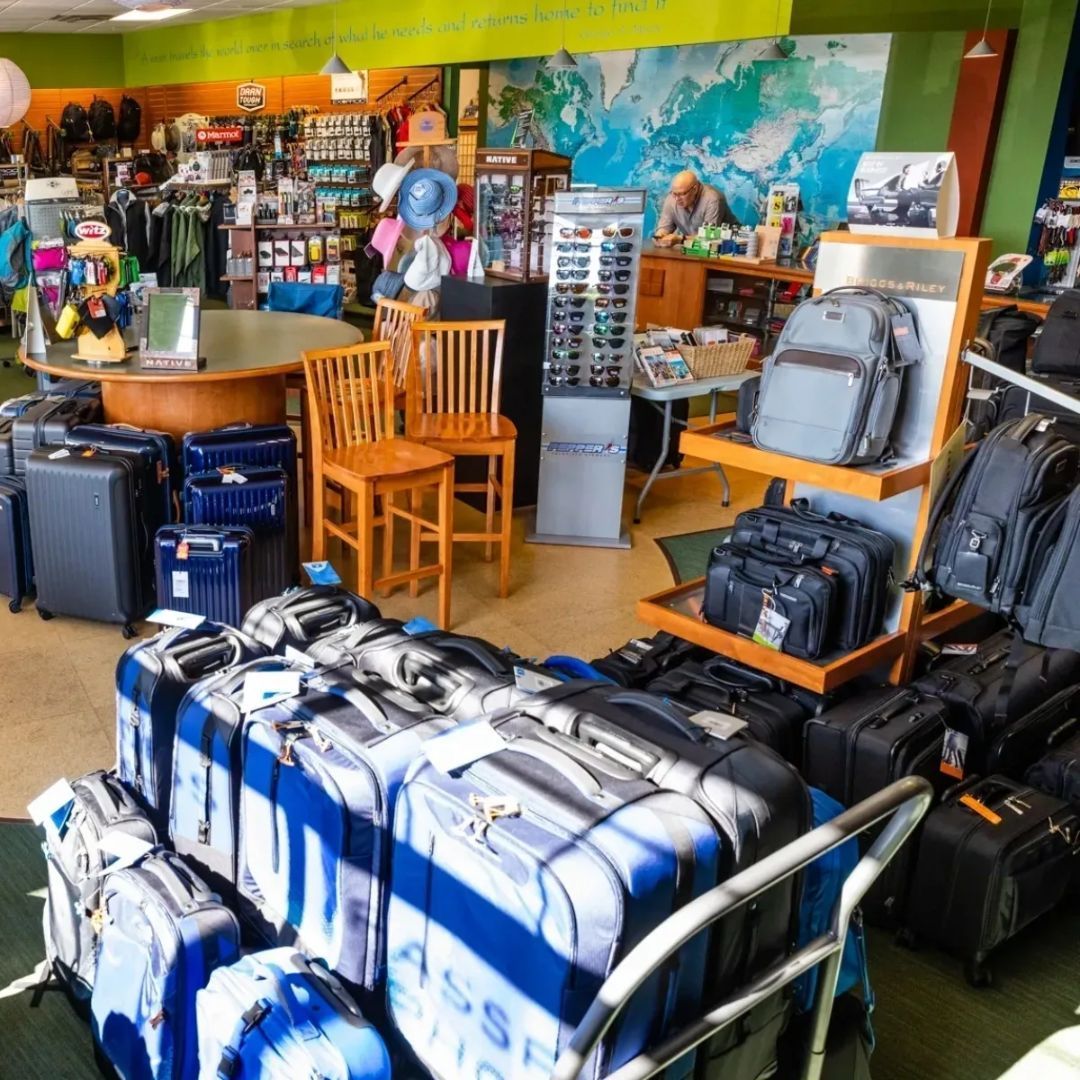 A retail travel store with rows of rolling luggage, suitcases, a display rack with bags, chairs, and a checkout counter.