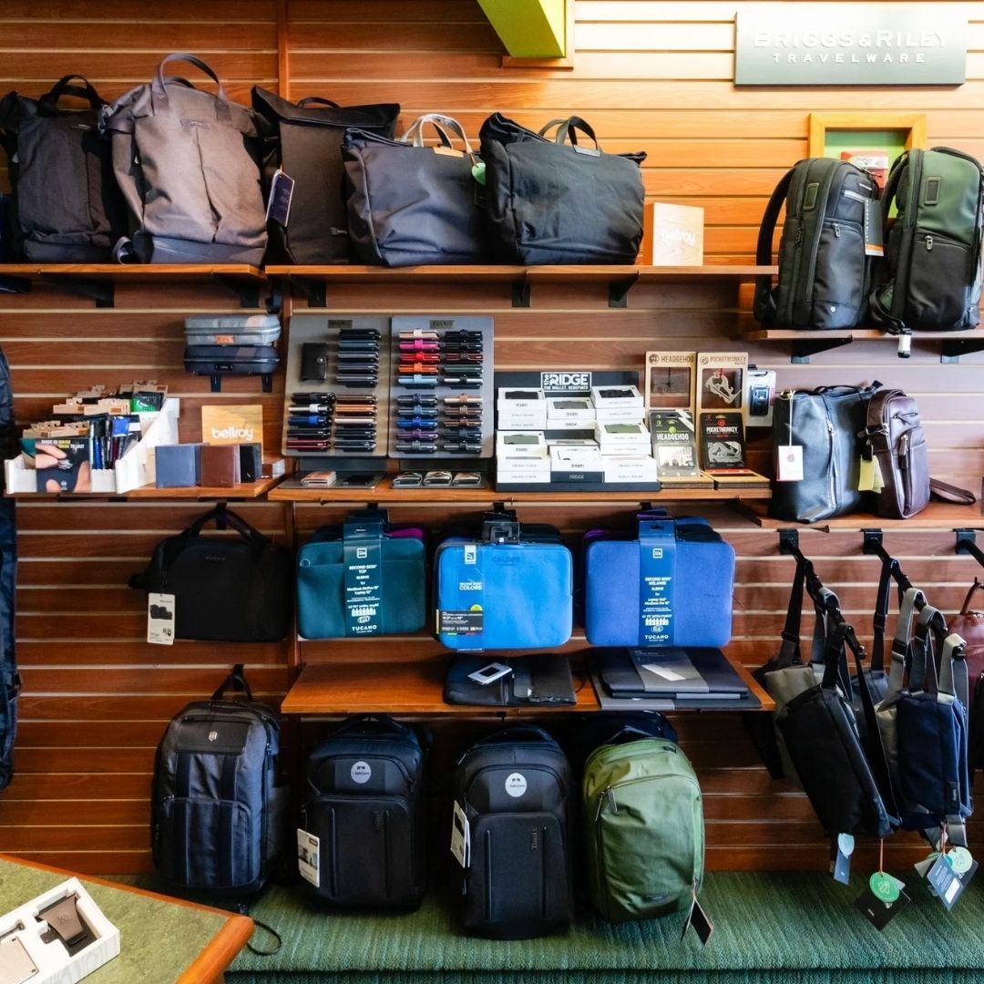 A retail store display of various backpacks, bags, and wallets arranged on wooden shelves against a slat wall.