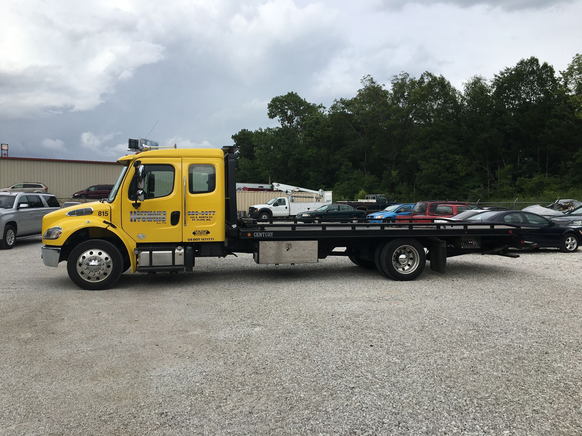 Yellow tow truck with a flatbed carrying several cars parked on a gravel lot under a cloudy sky.