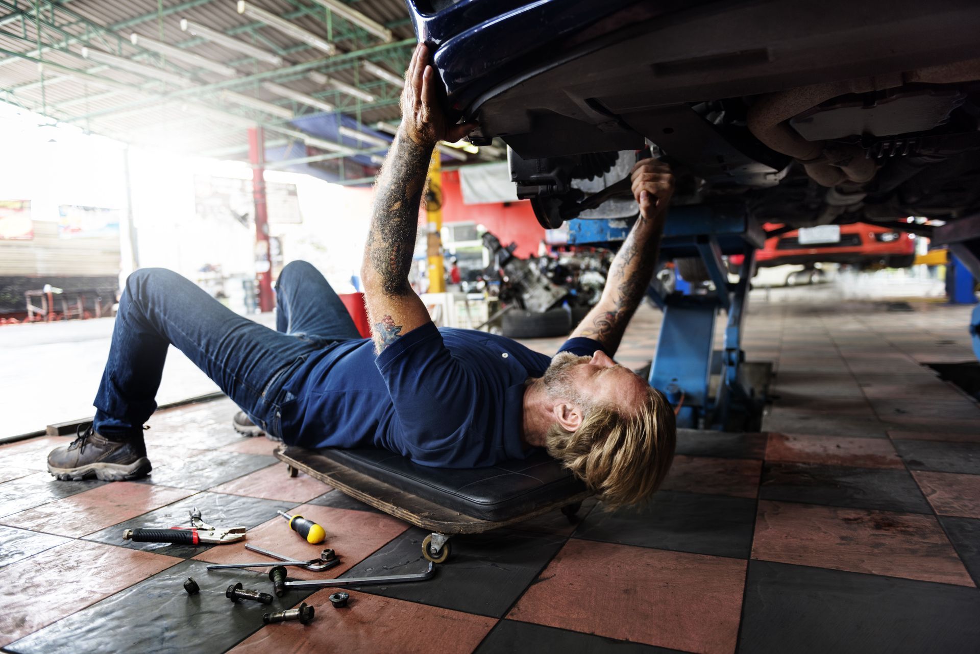 Mechanic working under a car in a repair shop. He is lying on a creeper and holding tools.