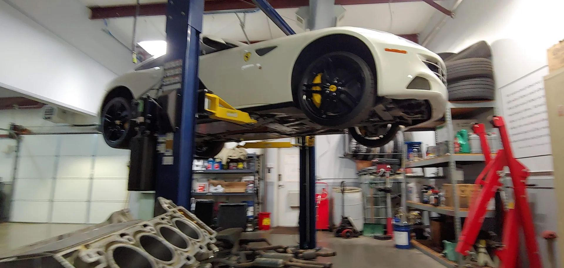 A white car is raised on a lift in a repair shop. Yellow calipers are visible. An engine block sits in the foreground.