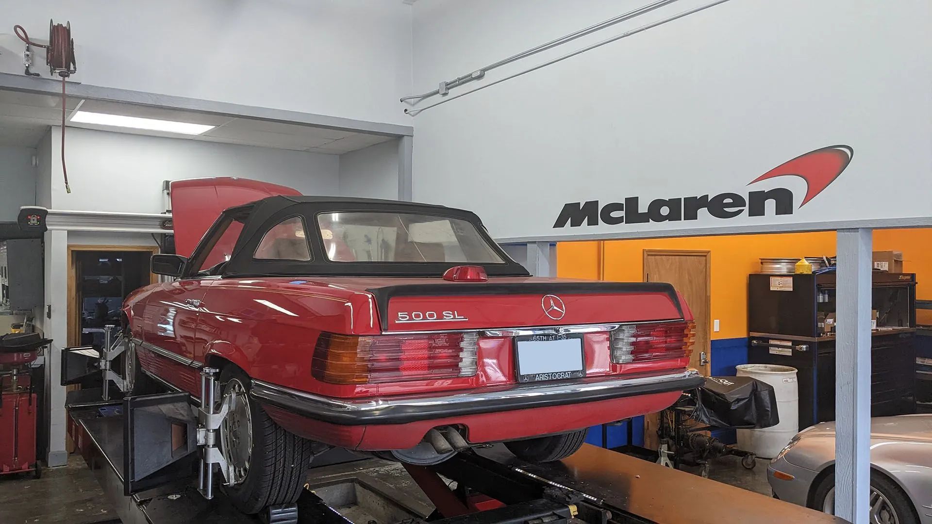 Red Mercedes convertible on a lift in a shop with a McLaren logo on the wall.