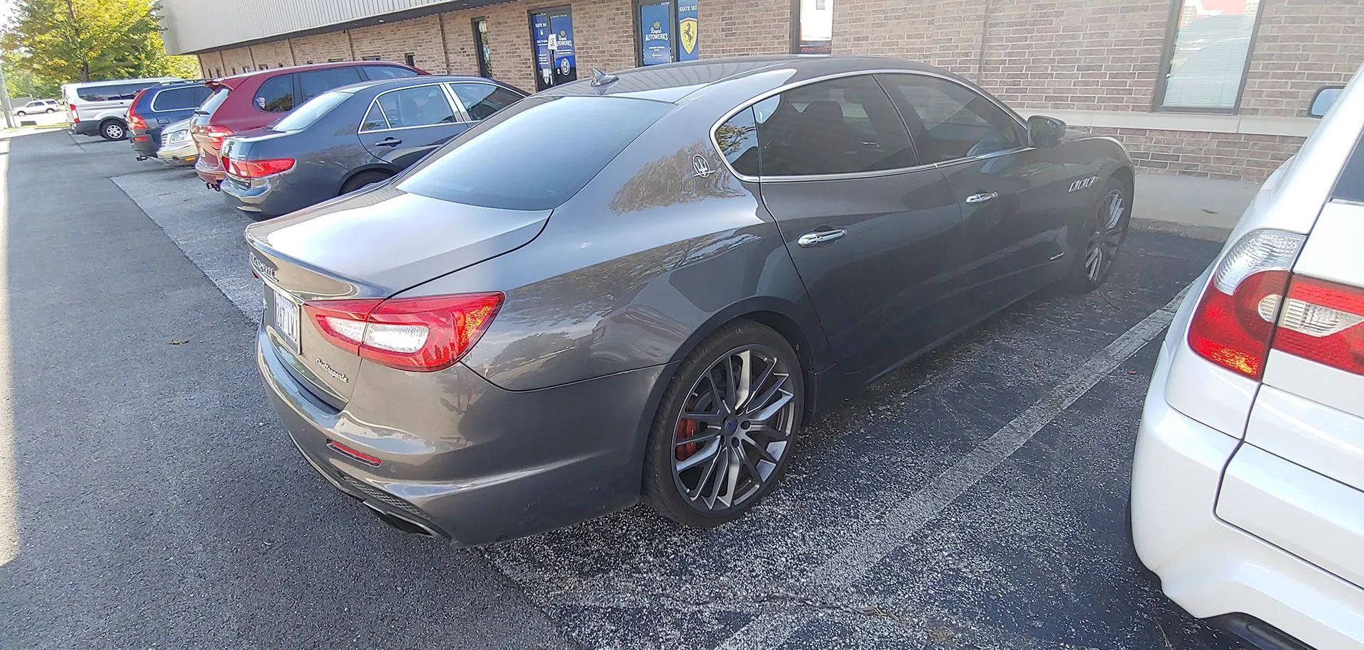 Gray Maserati sedan parked on gravel next to a white car. Other cars and a building are in the background.