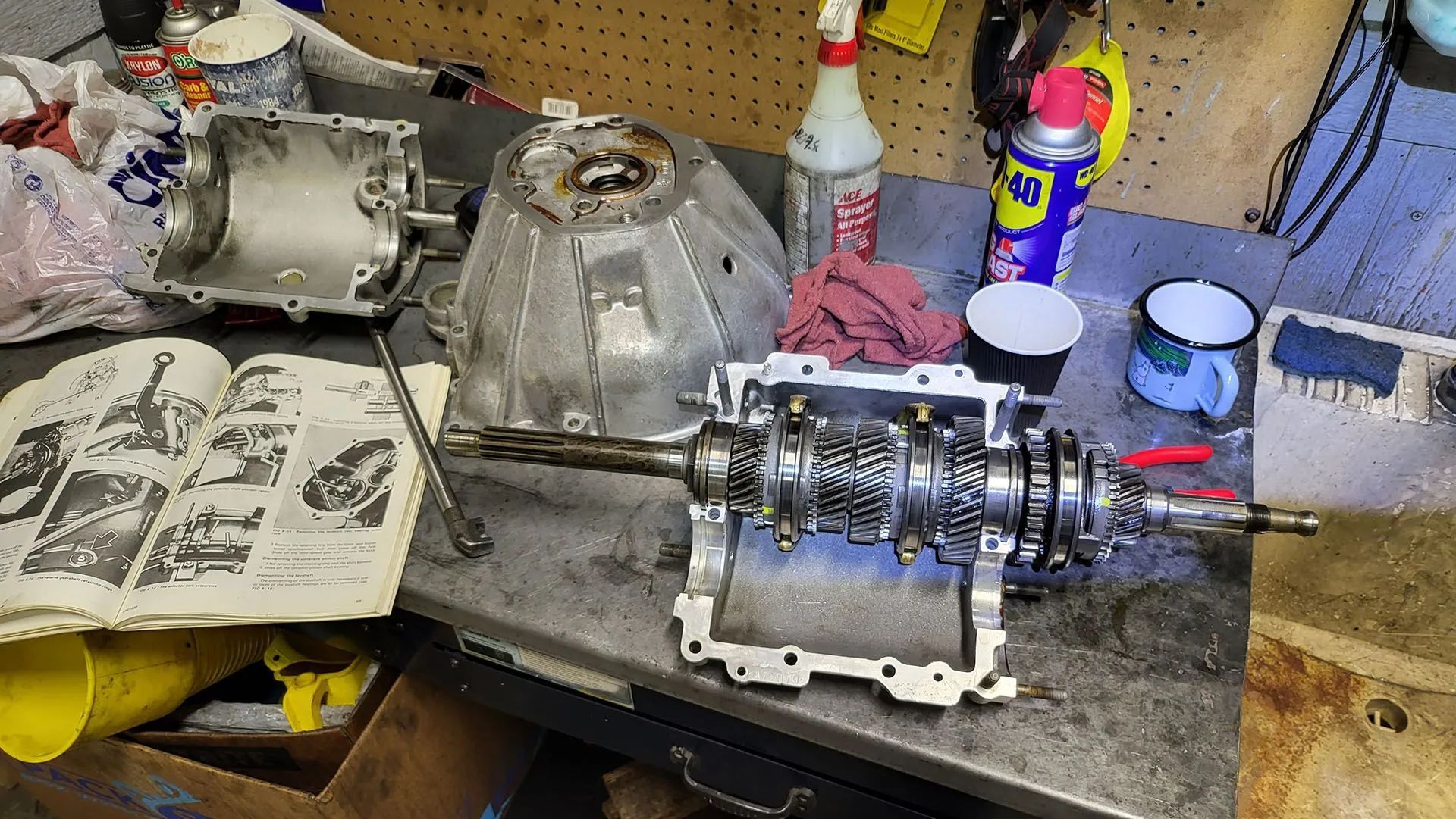 Disassembled transmission parts on a workbench with a repair manual. Silver, gray, and yellow items are present.