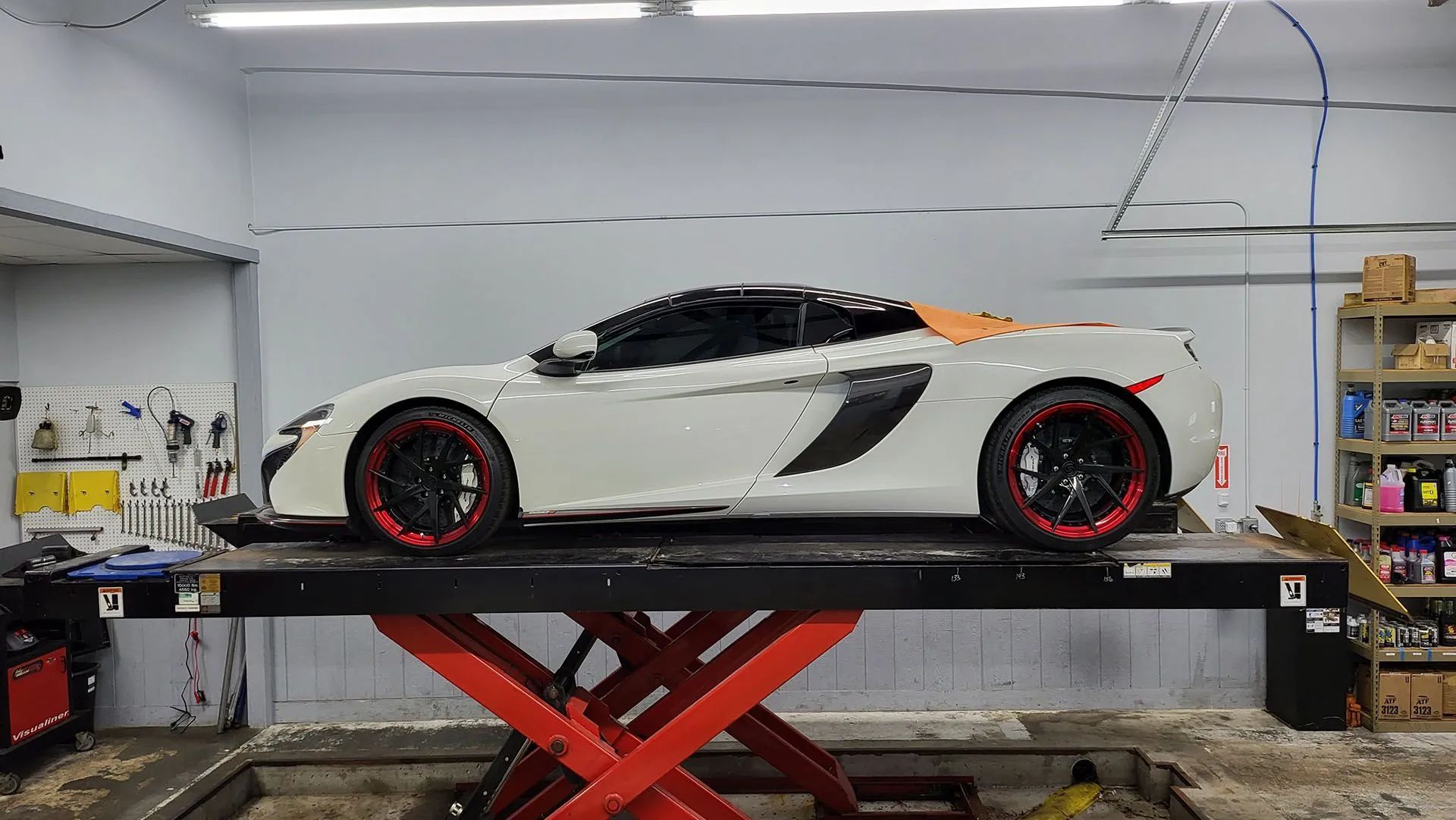 White sports car on a red scissor lift inside a workshop; black, orange, and red accents.
