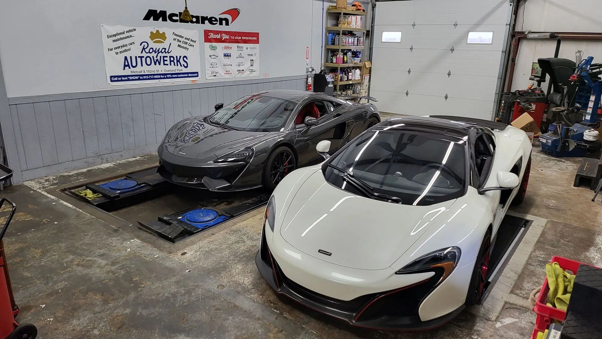 Two McLaren sports cars inside a garage: one gray, one white, with garage tools visible.