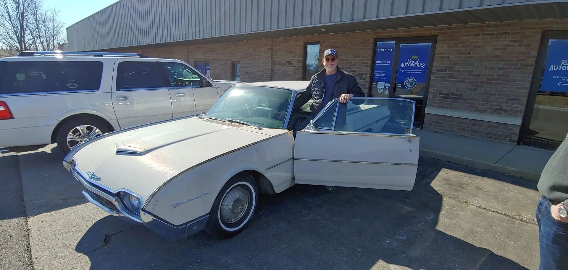A person stands next to an open-doored white Avanti car. Another SUV is parked next to them. Building in the background.