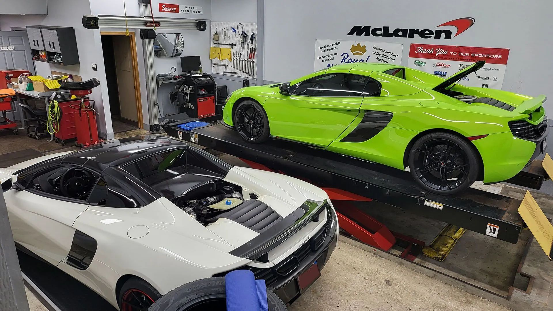 Two McLaren sports cars in a garage. One white, one neon green, on a lift. McLaren logo visible.