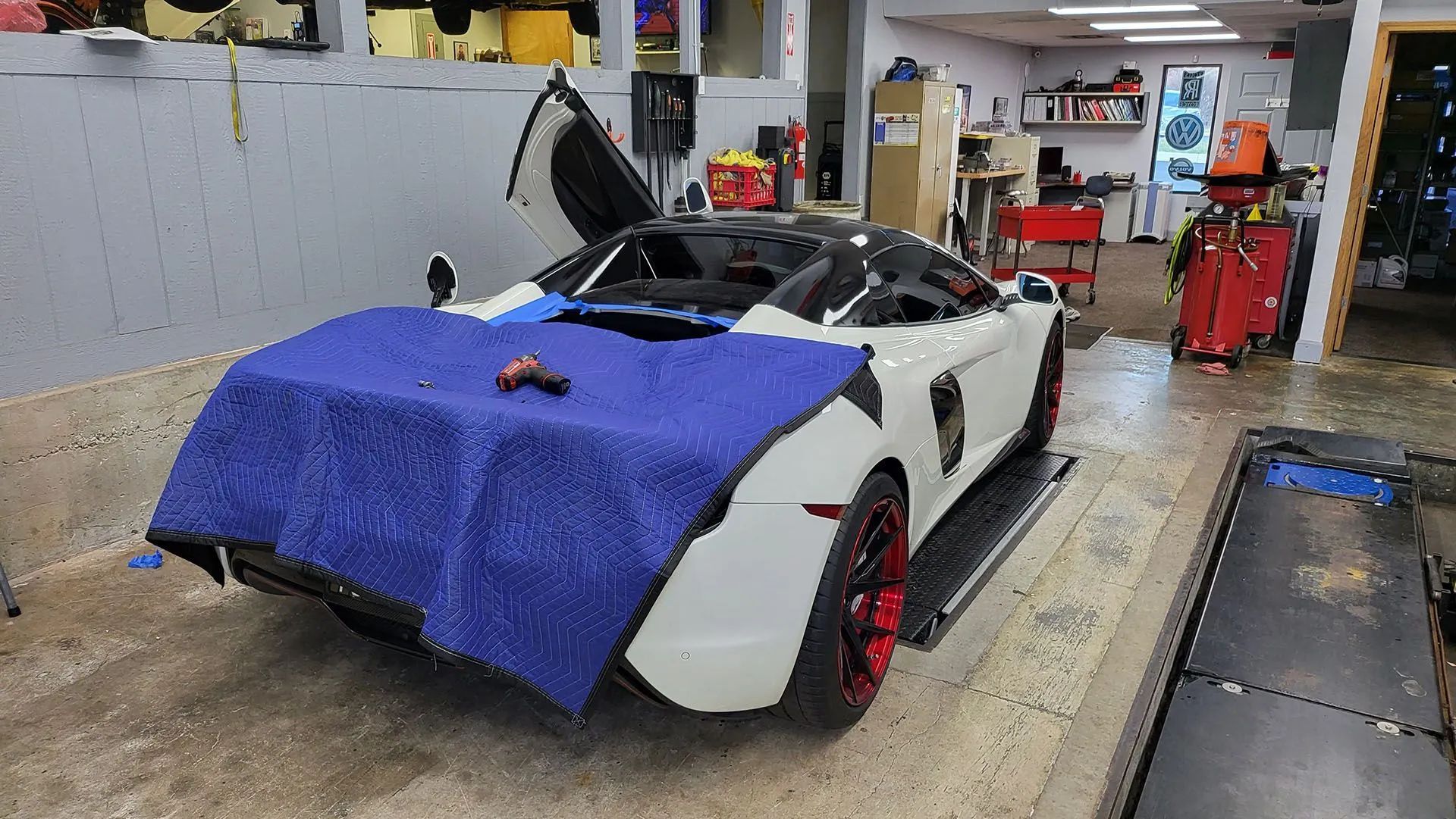 White sports car with open doors in a garage, draped with blue cover. Red rims.