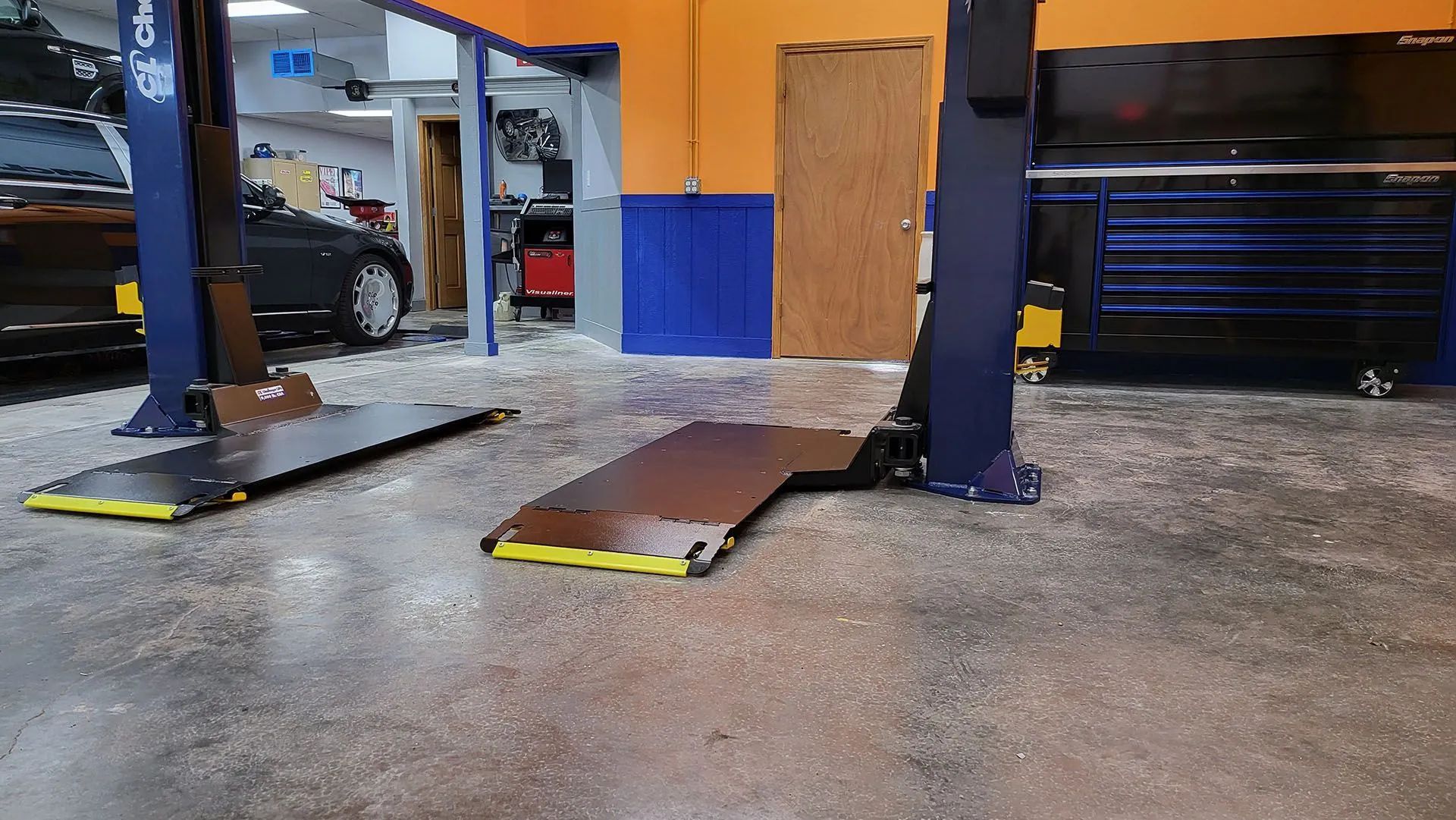 Two-post car lift in a garage with a car partially visible on the left. The floor is speckled.