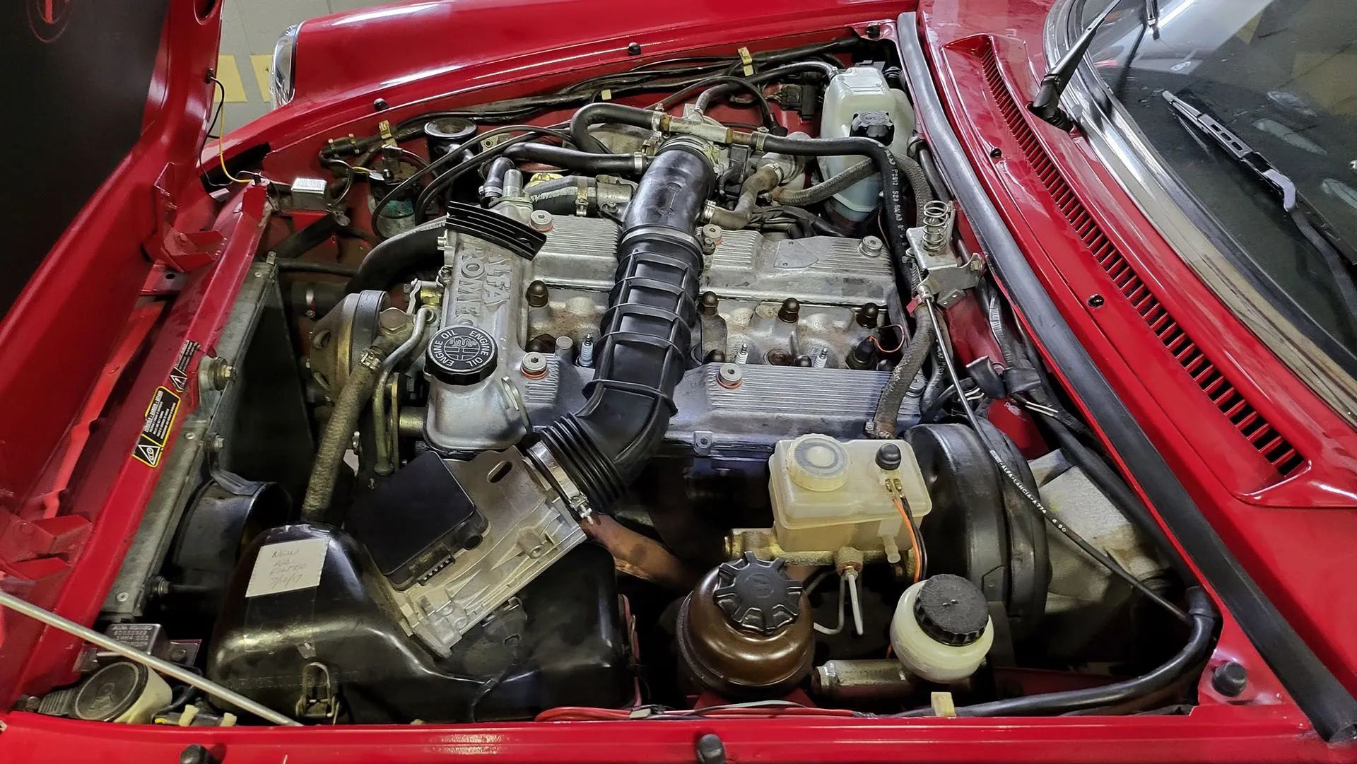Red car engine bay, open hood. Silver engine, black air intake, various hoses and containers.