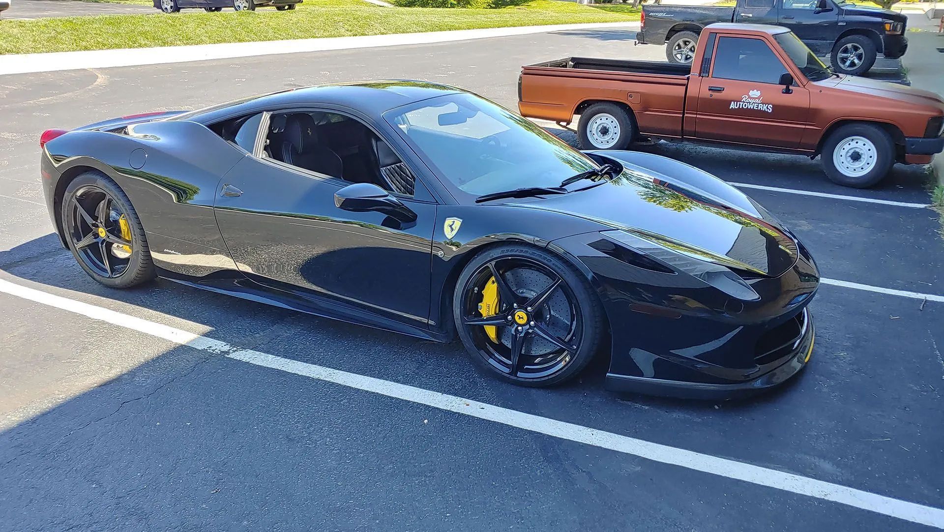 Black Ferrari sports car parked next to a brown pickup truck. Yellow brake calipers, outdoor setting.