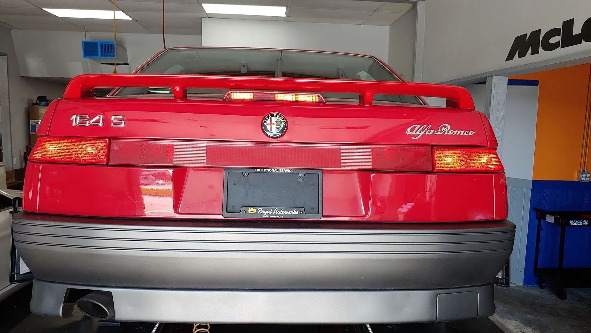 Red Alfa Romeo 164S rear view in a garage, gray bumper, black license plate, taillights and spoiler.