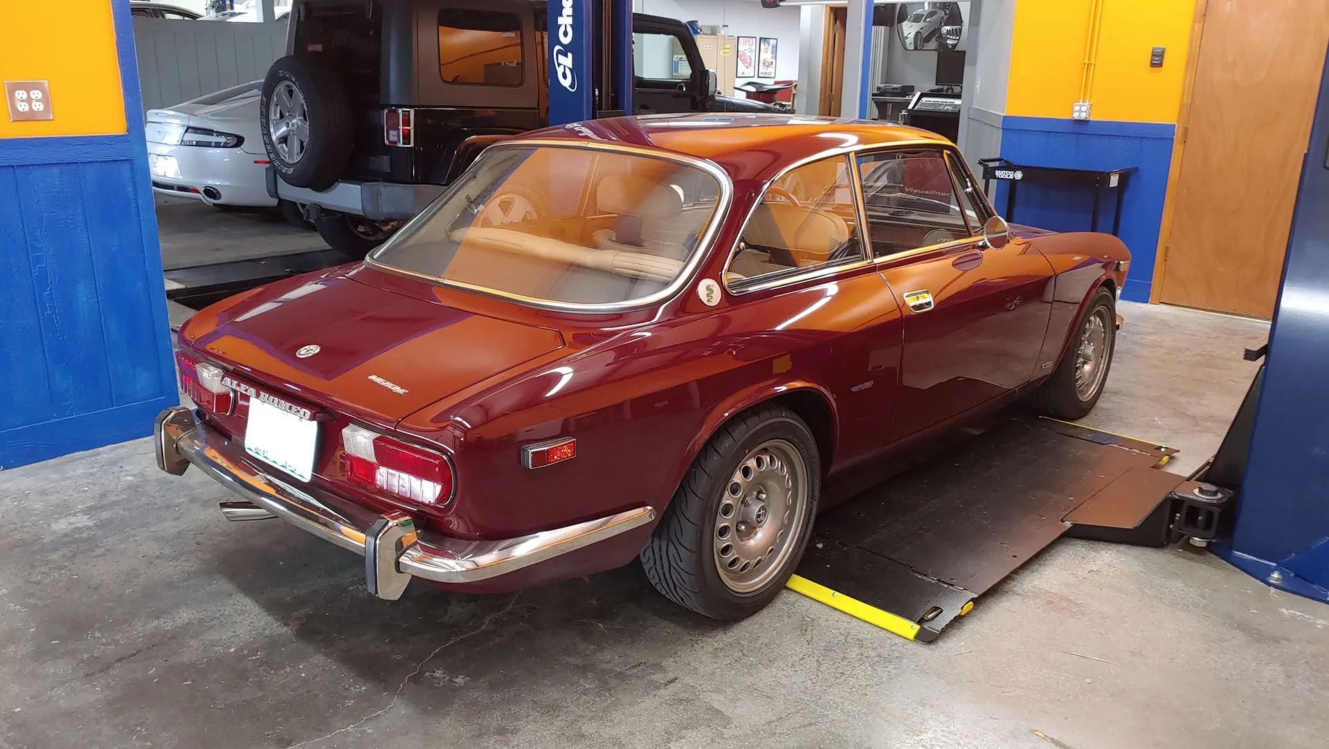 A vintage burgundy Alfa Romeo coupe in a garage, on a lift, with tan interior.