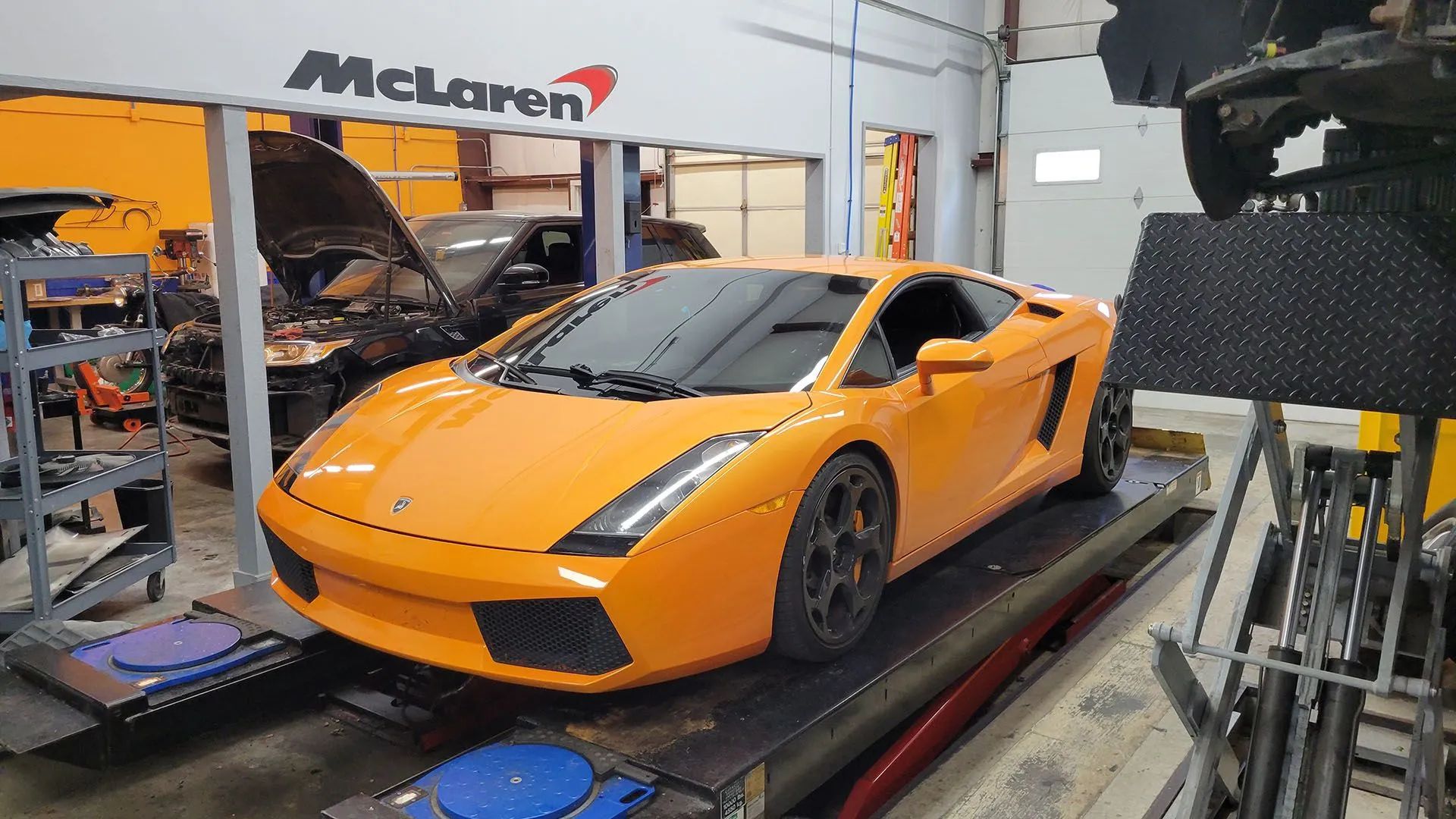 Orange Lamborghini on a lift in a garage with a McLaren sign above it.