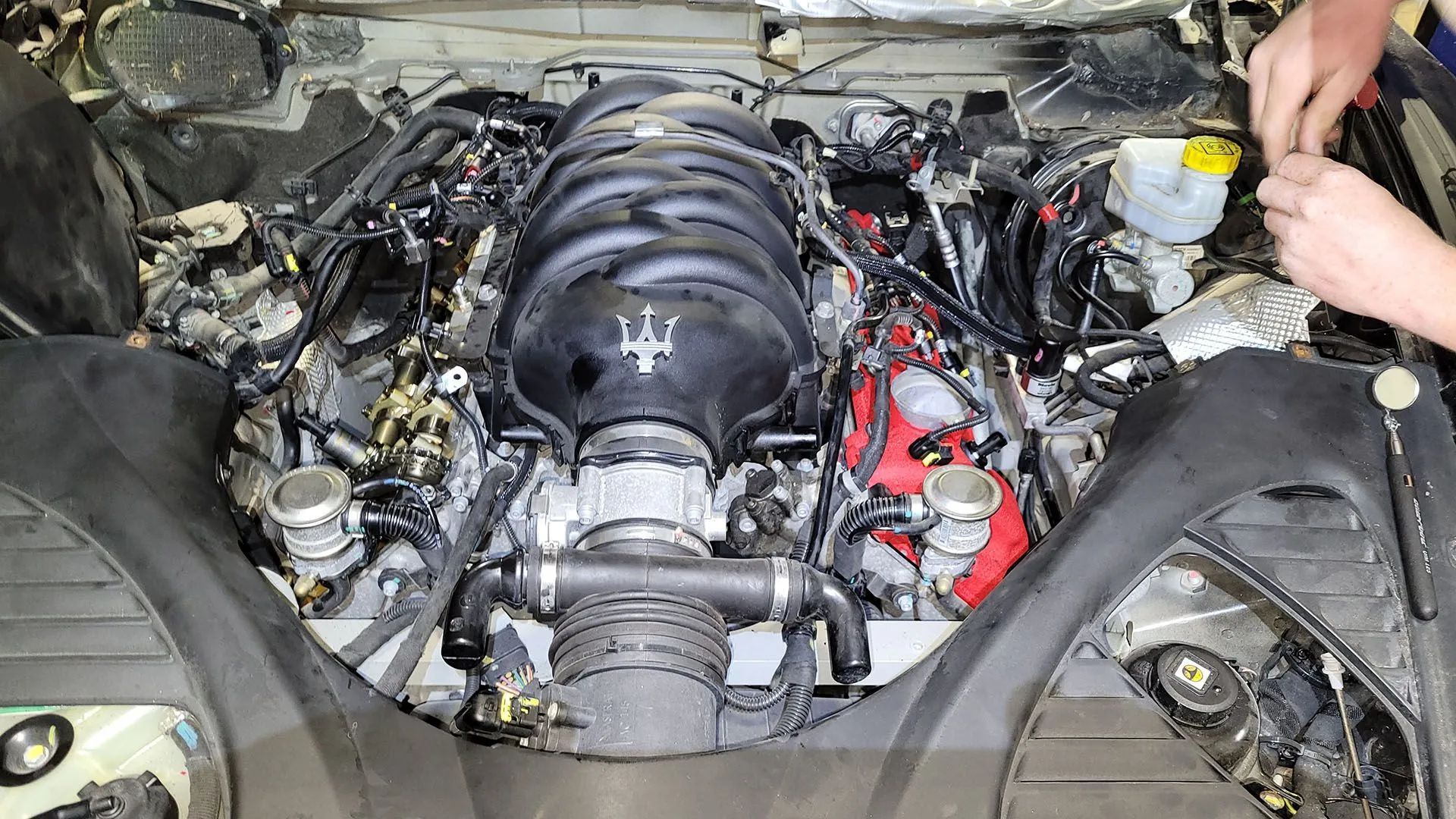 Engine of a Maserati with various components visible, mechanic working on it.