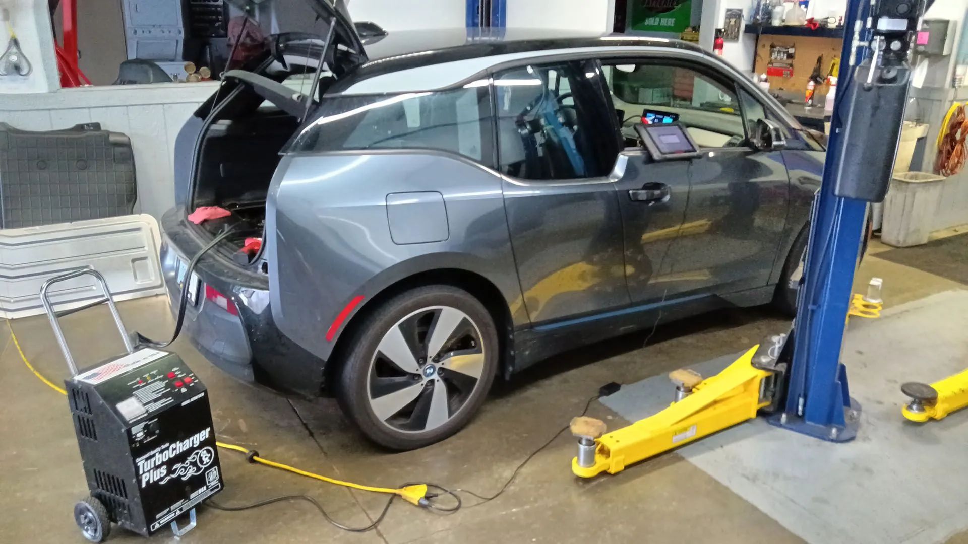 Gray electric car in a repair shop, open trunk, connected to a battery charger. Yellow lift.