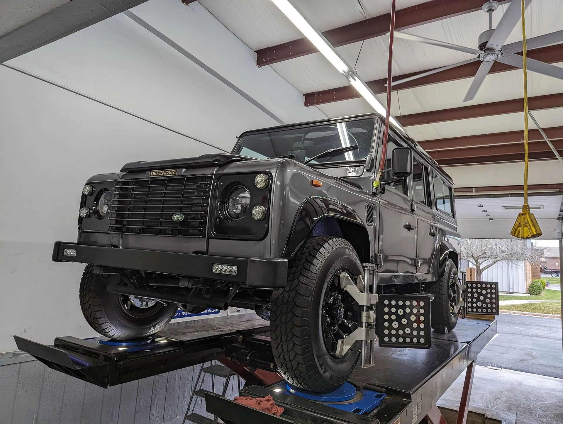 Gray Land Rover Defender on a lift in a garage, undergoing an alignment, with alignment tools on wheels.