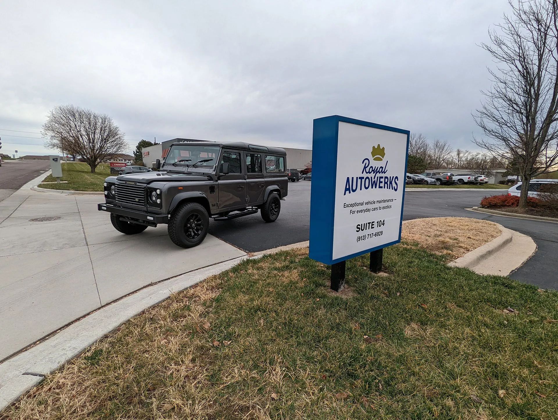 Dark gray Land Rover Defender parked by a sign for Royal AutoHaus on a cloudy day.