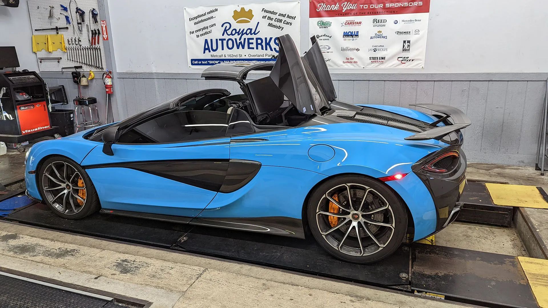 Blue McLaren convertible with doors up in a garage, orange brake calipers.
