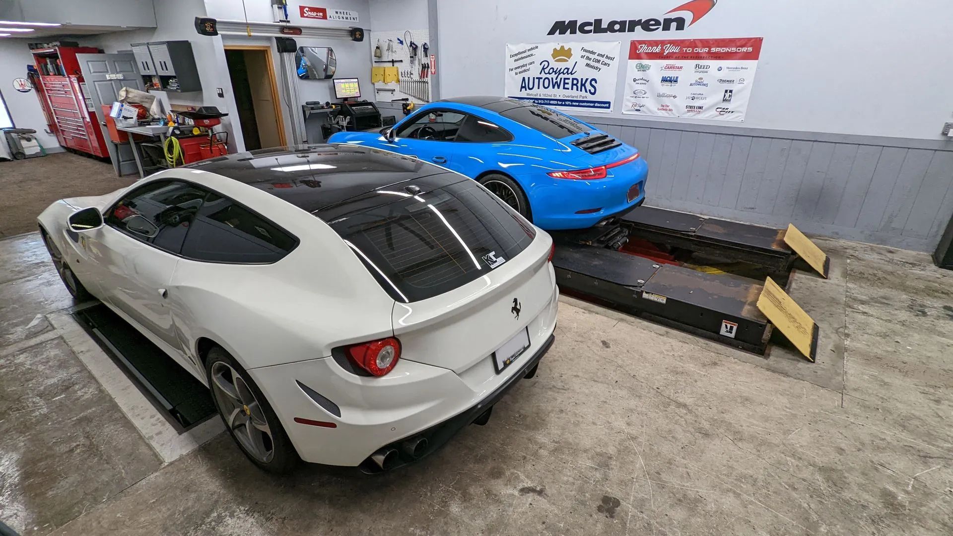 White Ferrari and blue Porsche inside a garage. Cars on lifts, working on engines.