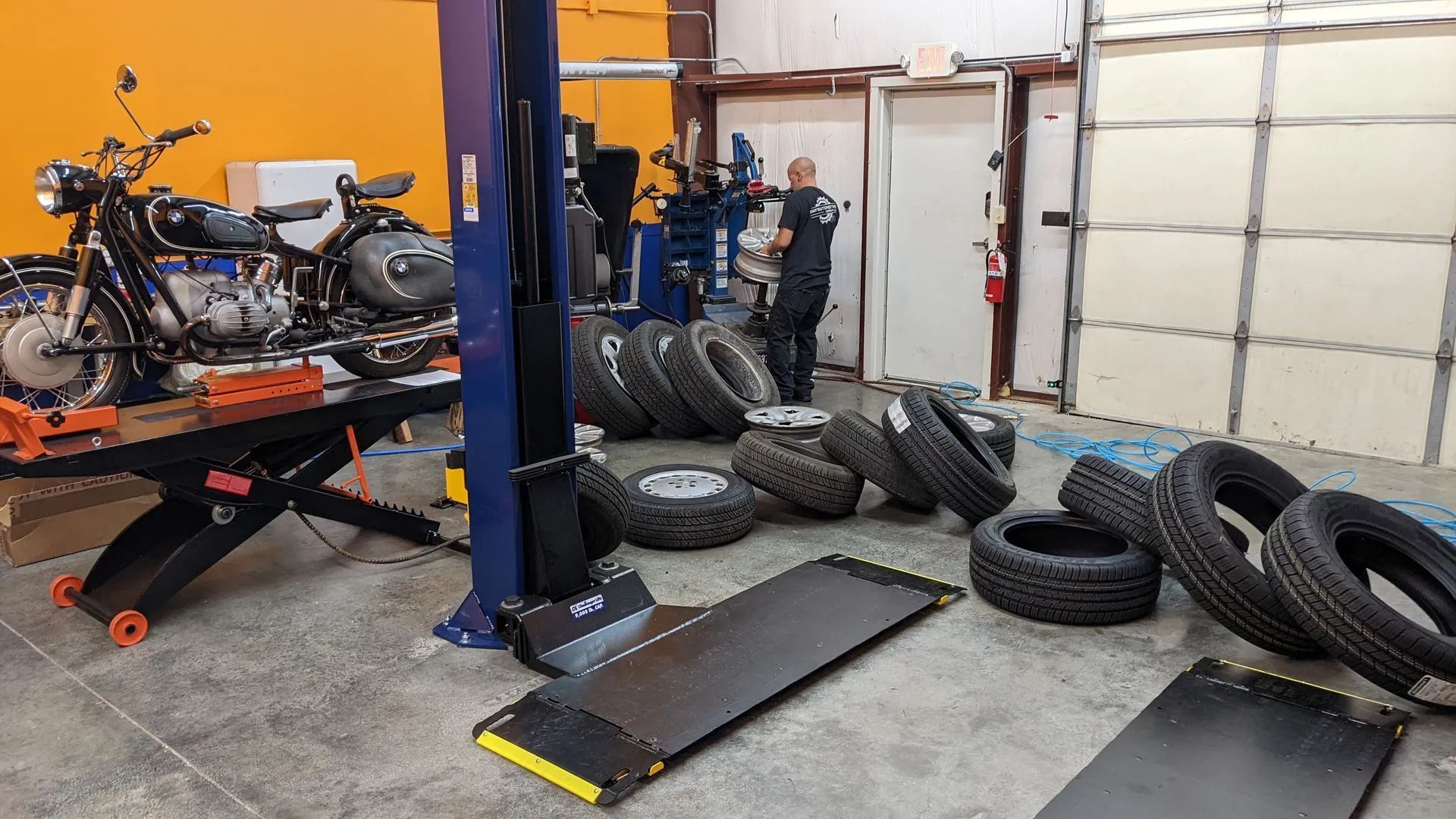 Motorcycle on lift in auto shop with tires, a mechanic, and equipment.