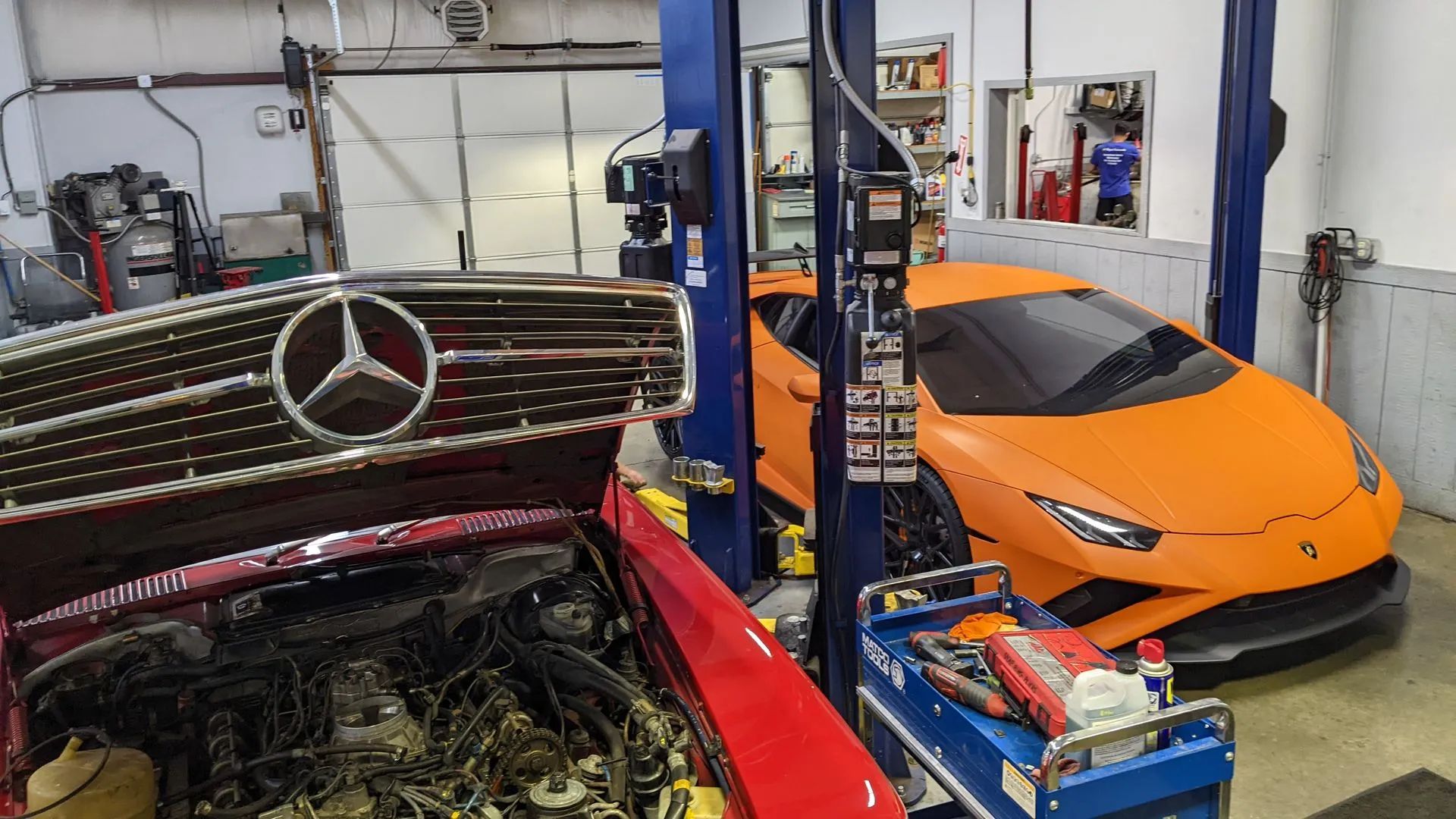 Red Mercedes with open hood and orange Lamborghini in a garage.