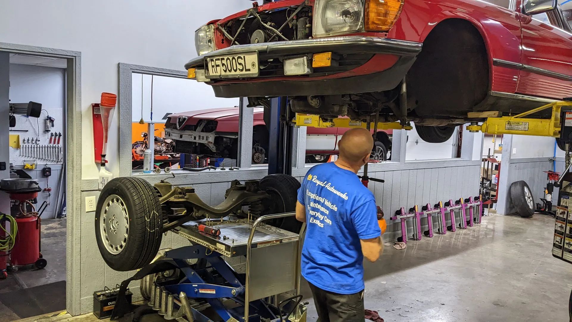 Man working on a red car lifted in a garage. Car's undercarriage exposed.  Tools and another car are visible in the background.
