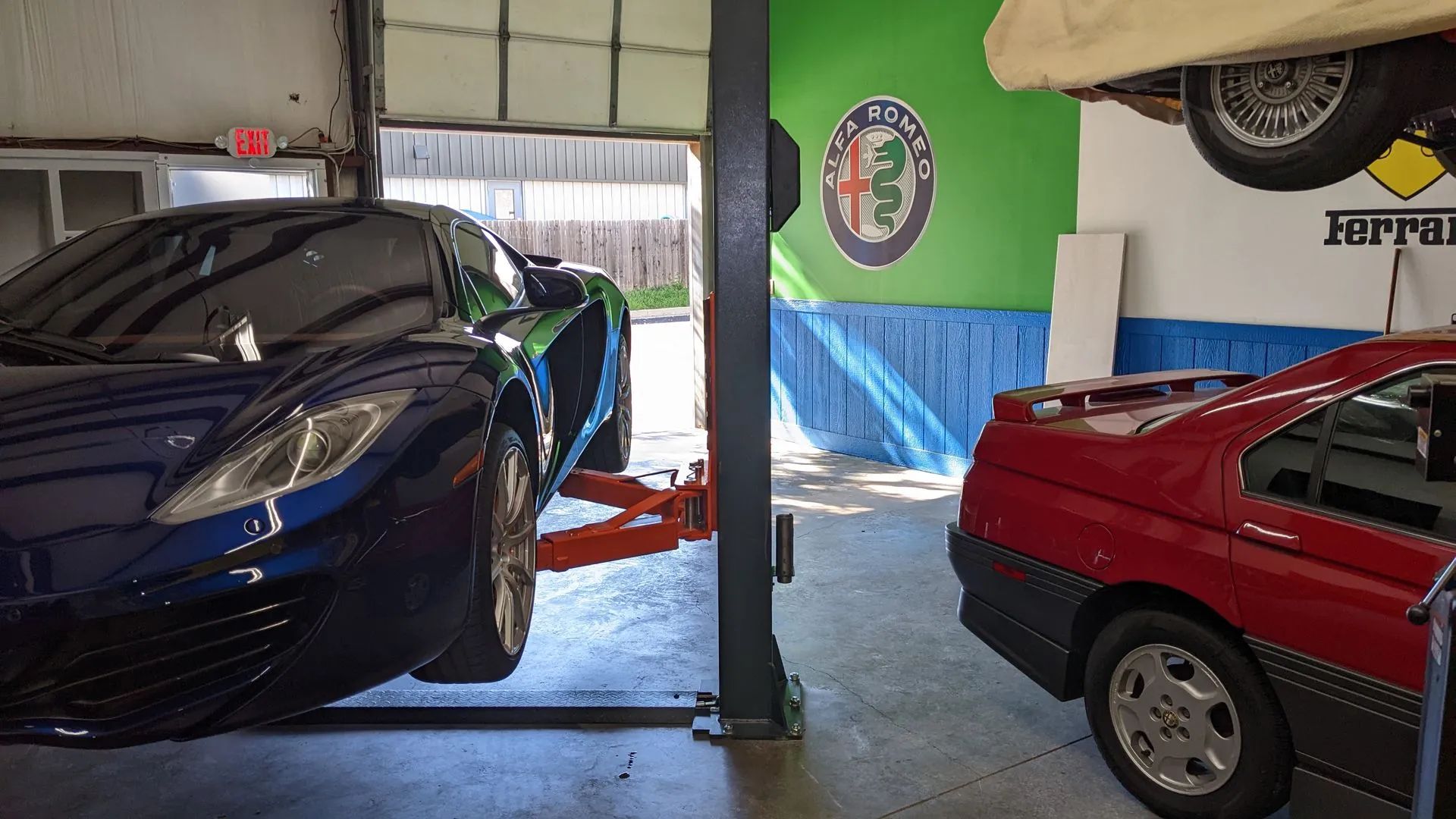 A blue sports car on a lift and a red car in a garage. Alfa Romeo logo on the wall.