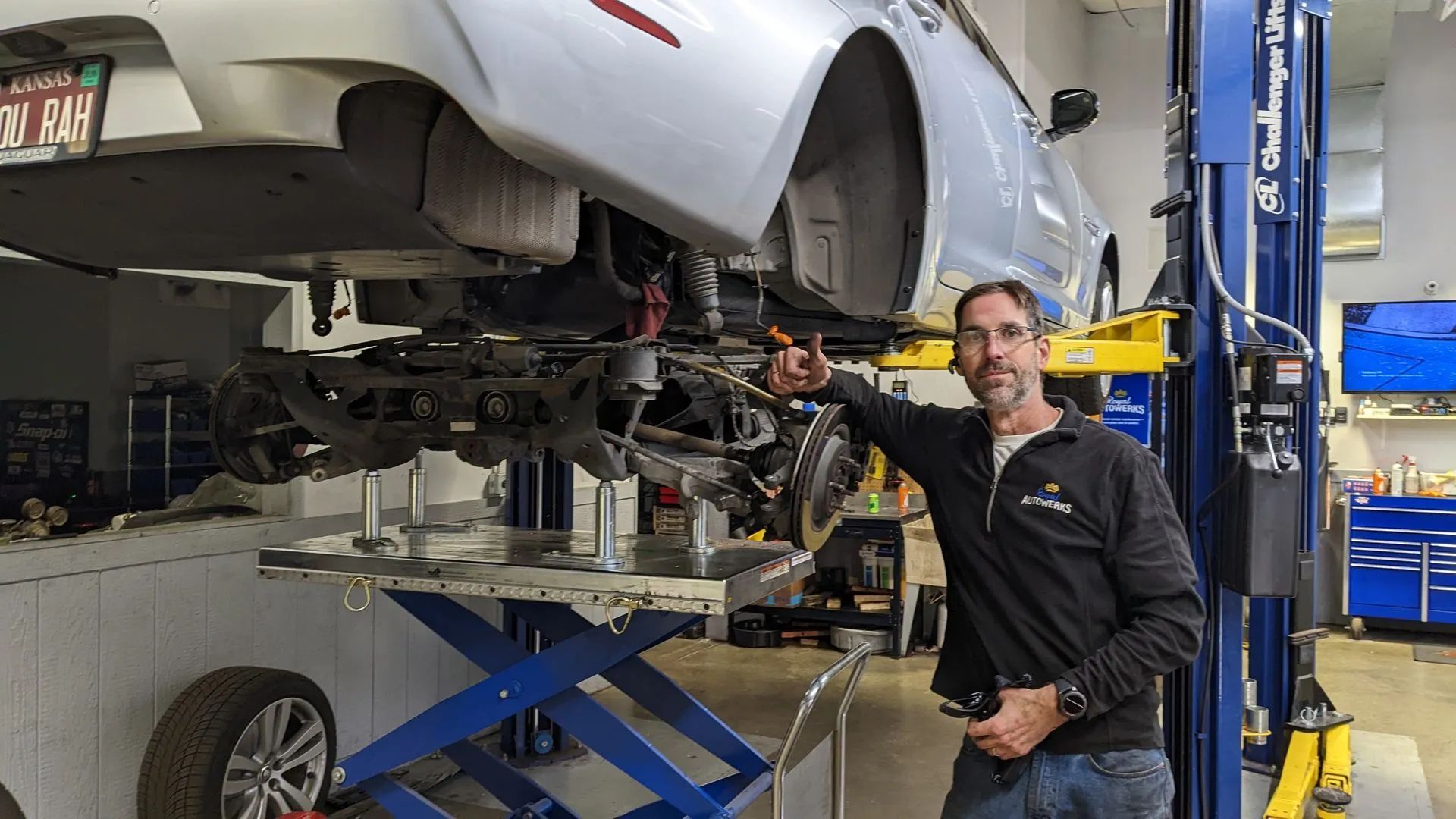 Mechanic beside a silver car lifted on a blue hydraulic platform in a shop; he is holding a tool.