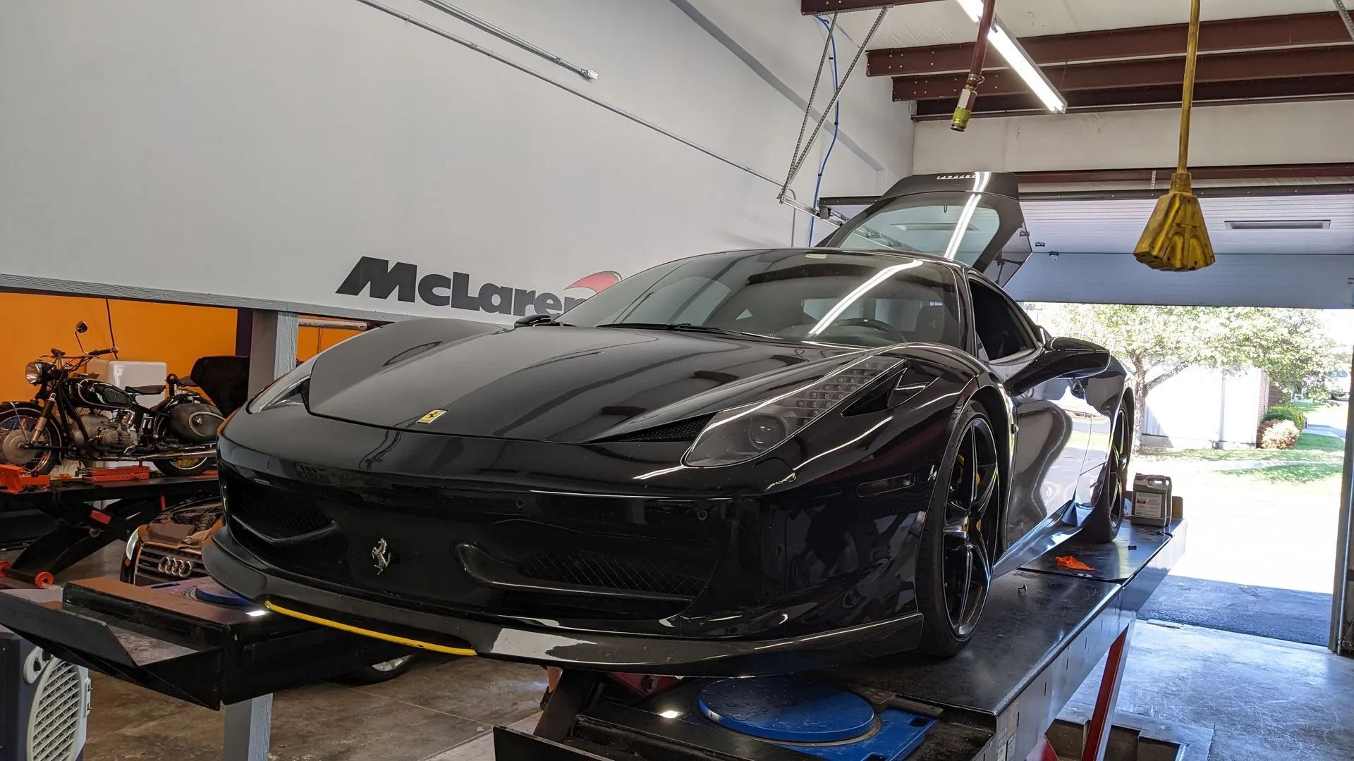 Black Ferrari sports car on a lift inside a garage. The door is open, and there is a McLaren logo on the wall.