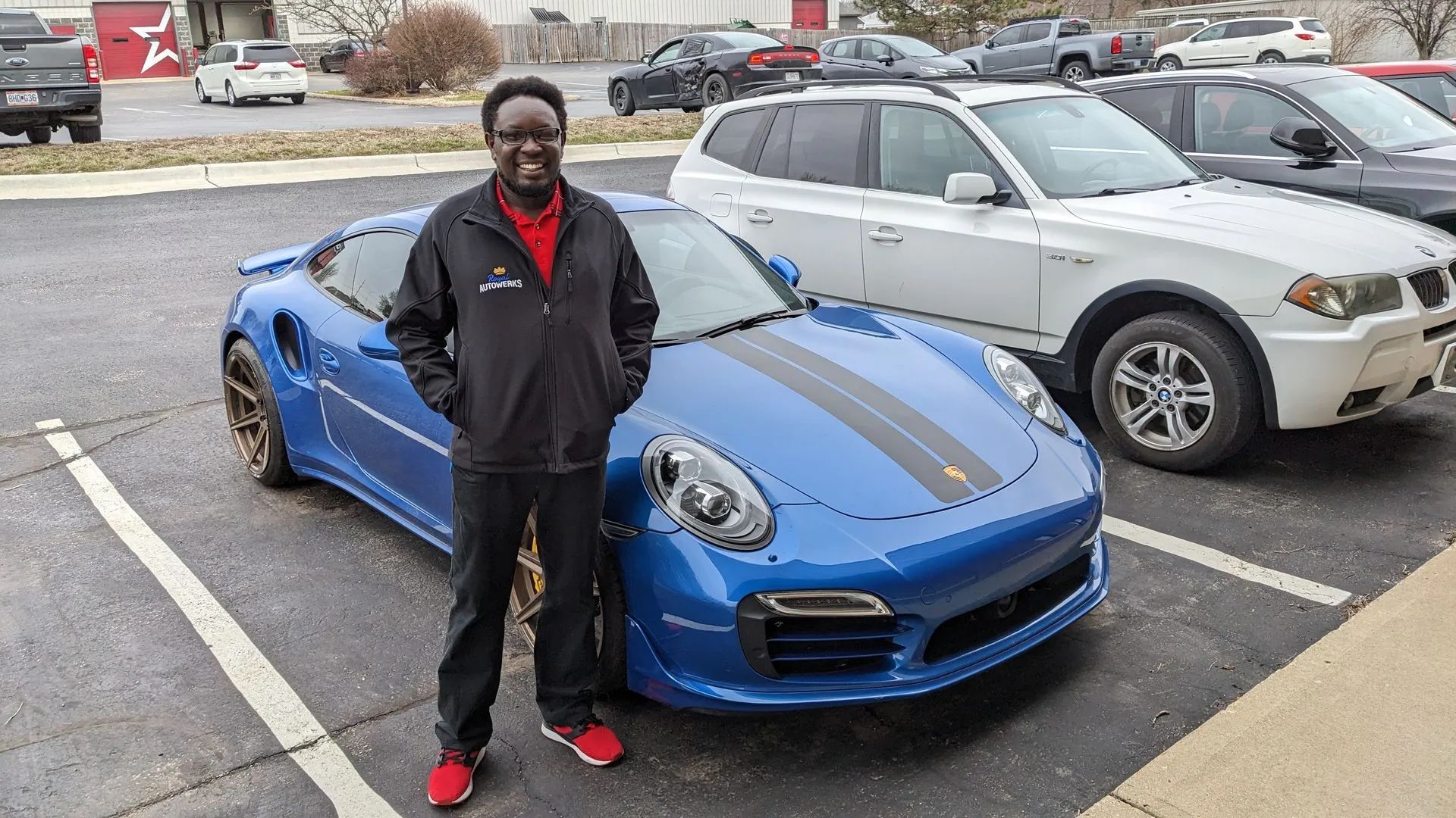 Man standing next to a blue Porsche sports car with black racing stripes in a parking lot.